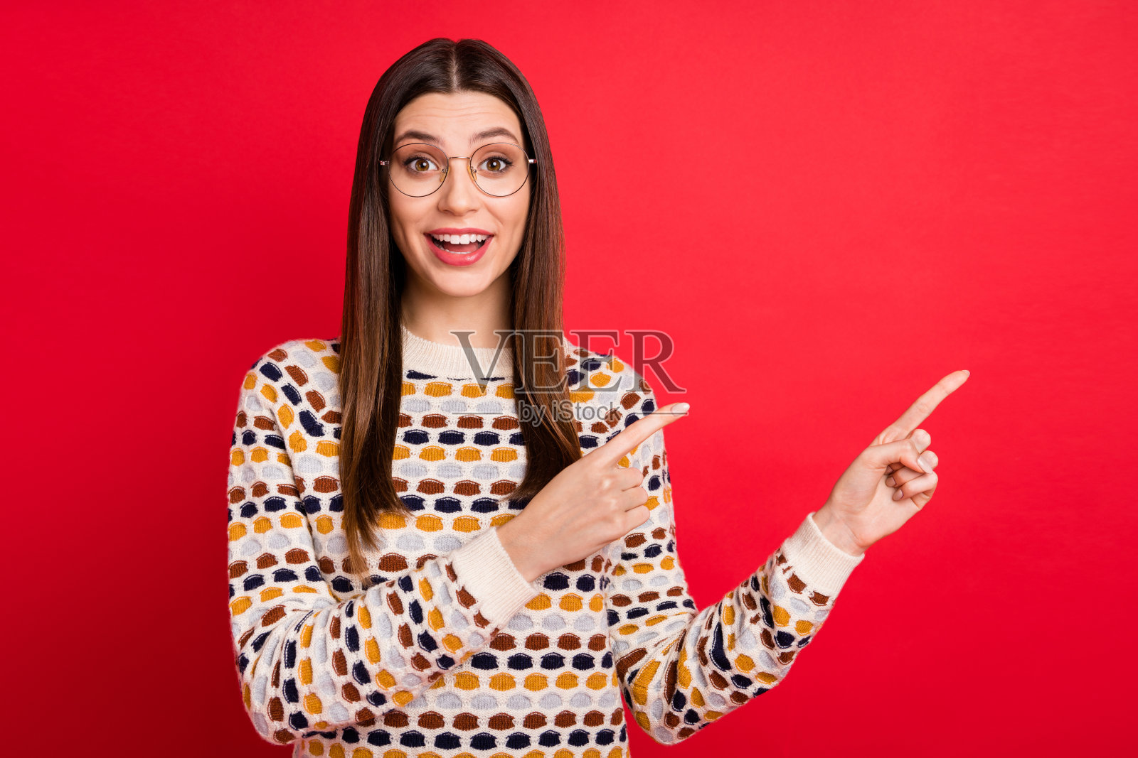 穿着彩色毛衣的可爱女孩，惊讶地指着空白处，背景为红色。照片摄影图片