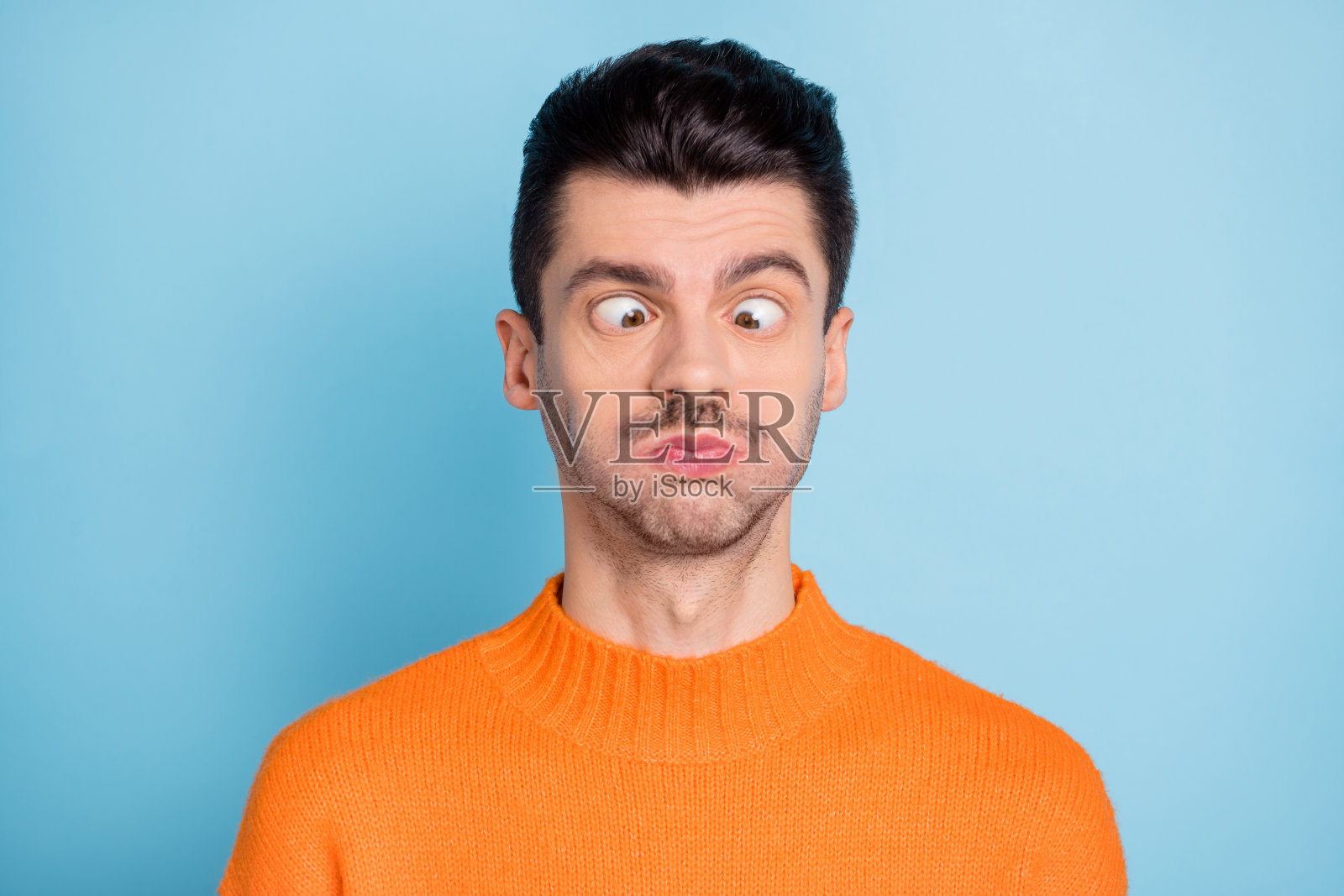 年轻男子搞怪傻乎乎地捂住嘴巴，单独站在蓝色背景前。照片摄影图片