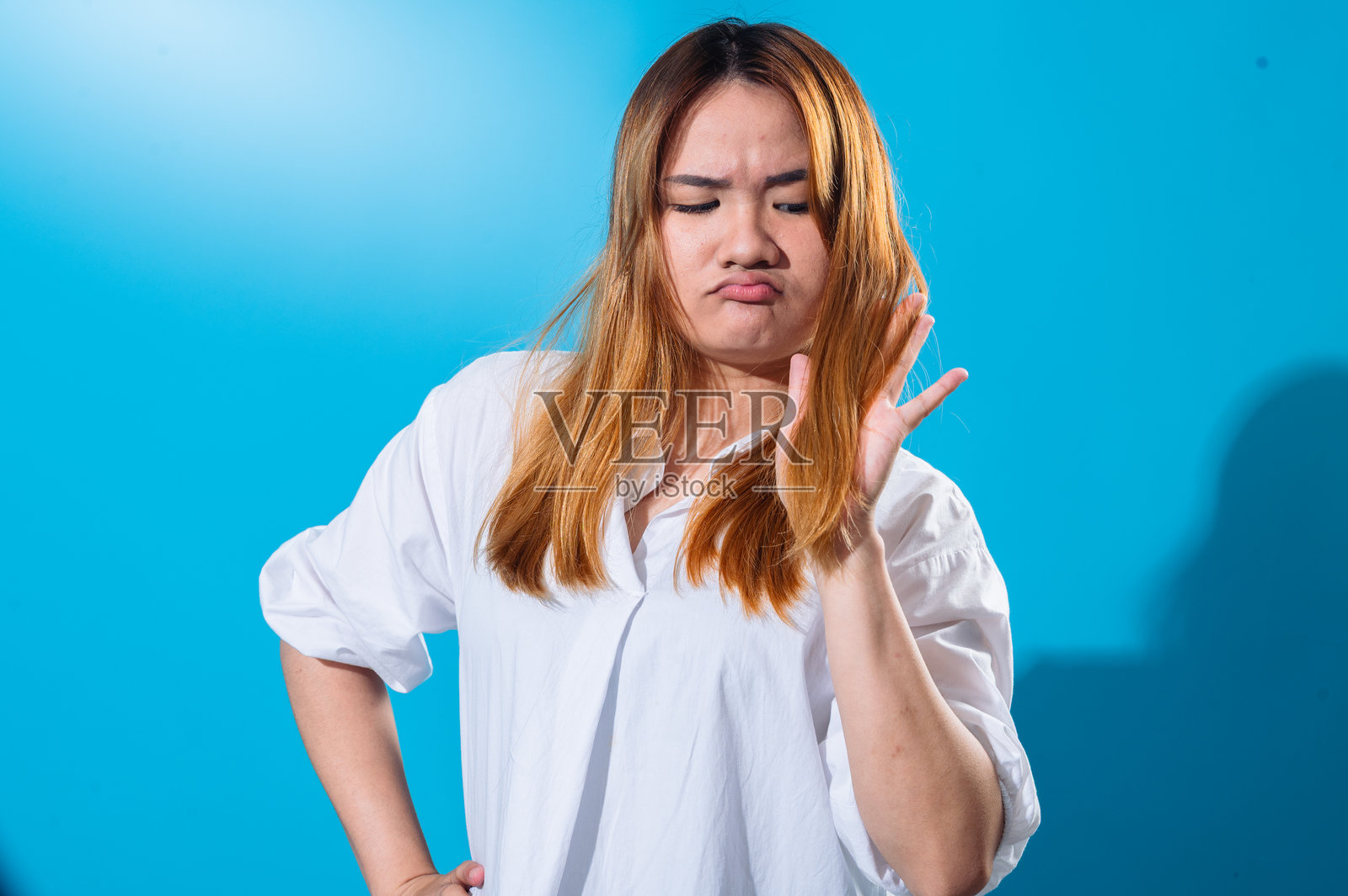 一位穿着白衬衫的亚洲女性，面露担忧和不满的表情，正在检查自己的头发，观察发梢分叉或干燥，背景为单色蓝色，灯光为摄影棚灯光。照片摄影图片