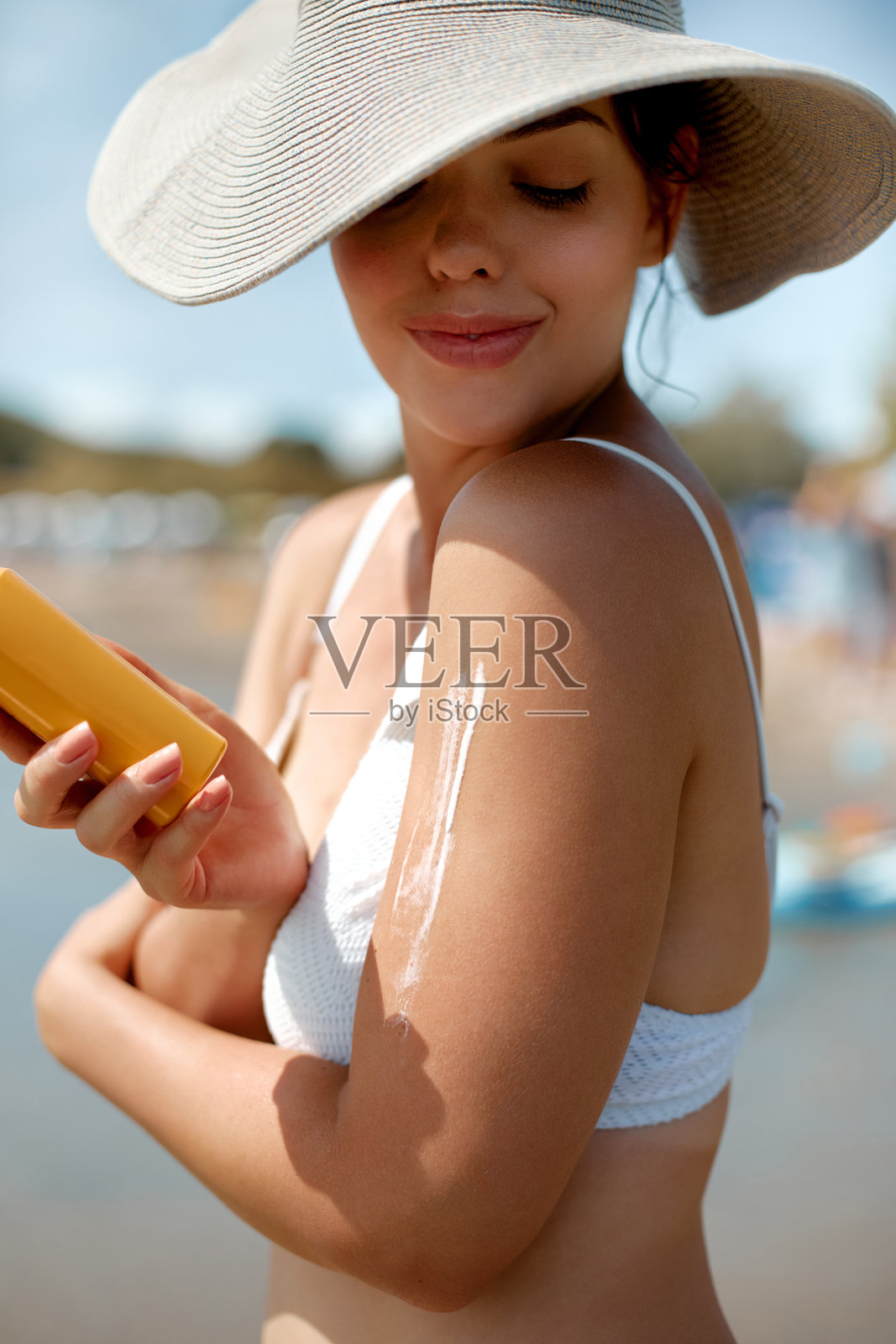 女性在海滩上戴着宽边帽涂抹防晒霜以保护皮肤照片摄影图片
