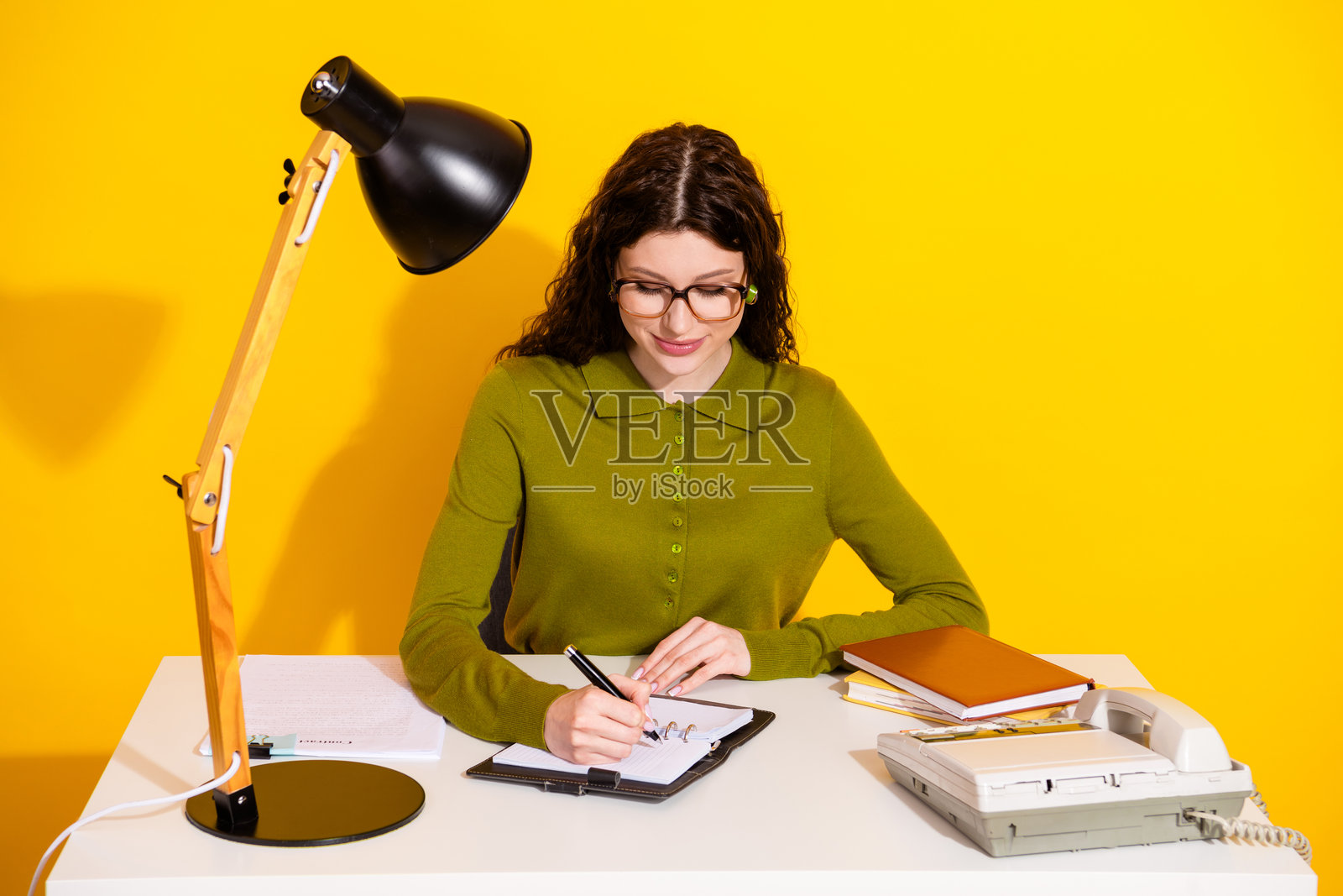 年轻女性坐在桌子前，穿着时尚的卡其色衬衫，在黄色背景下用复古道具写日记。照片摄影图片