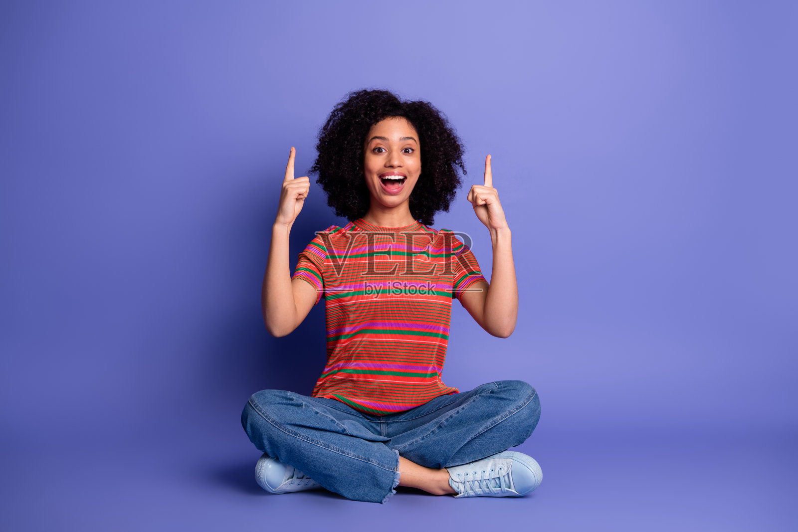 快乐的年轻女性盘腿而坐，指向上方，背景为生动的紫色，展现出喜悦和兴奋。照片摄影图片