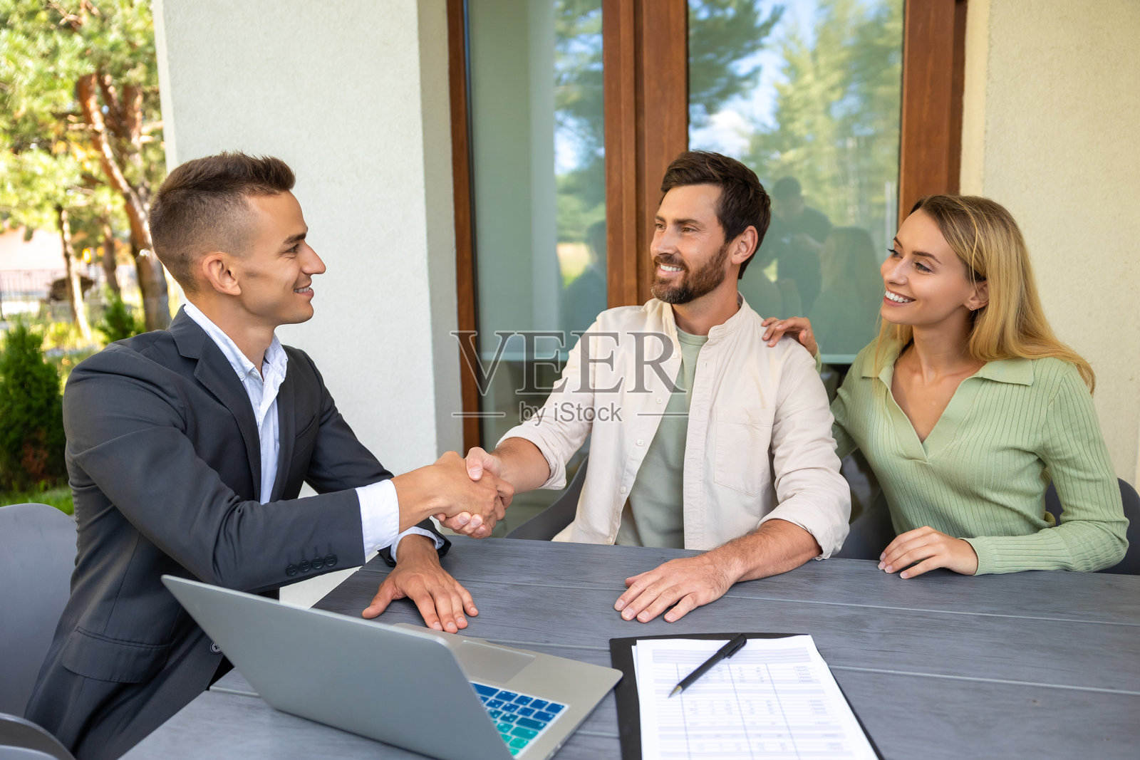 房地产经纪人与一对夫妇在户外桌子上签署房产交易协议，桌上有笔记本电脑和文件。照片摄影图片