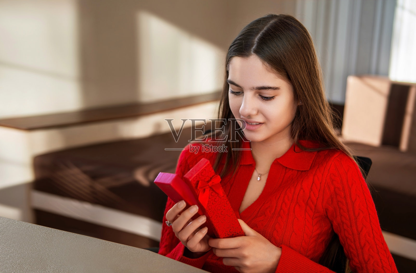 年轻女性在室内打开一个红色礼品盒。她穿着红色毛衣，手里拿着并打开红色礼品盒，脸上流露出好奇和快乐的表情，周围是一个温馨的房间环境。照片摄影图片