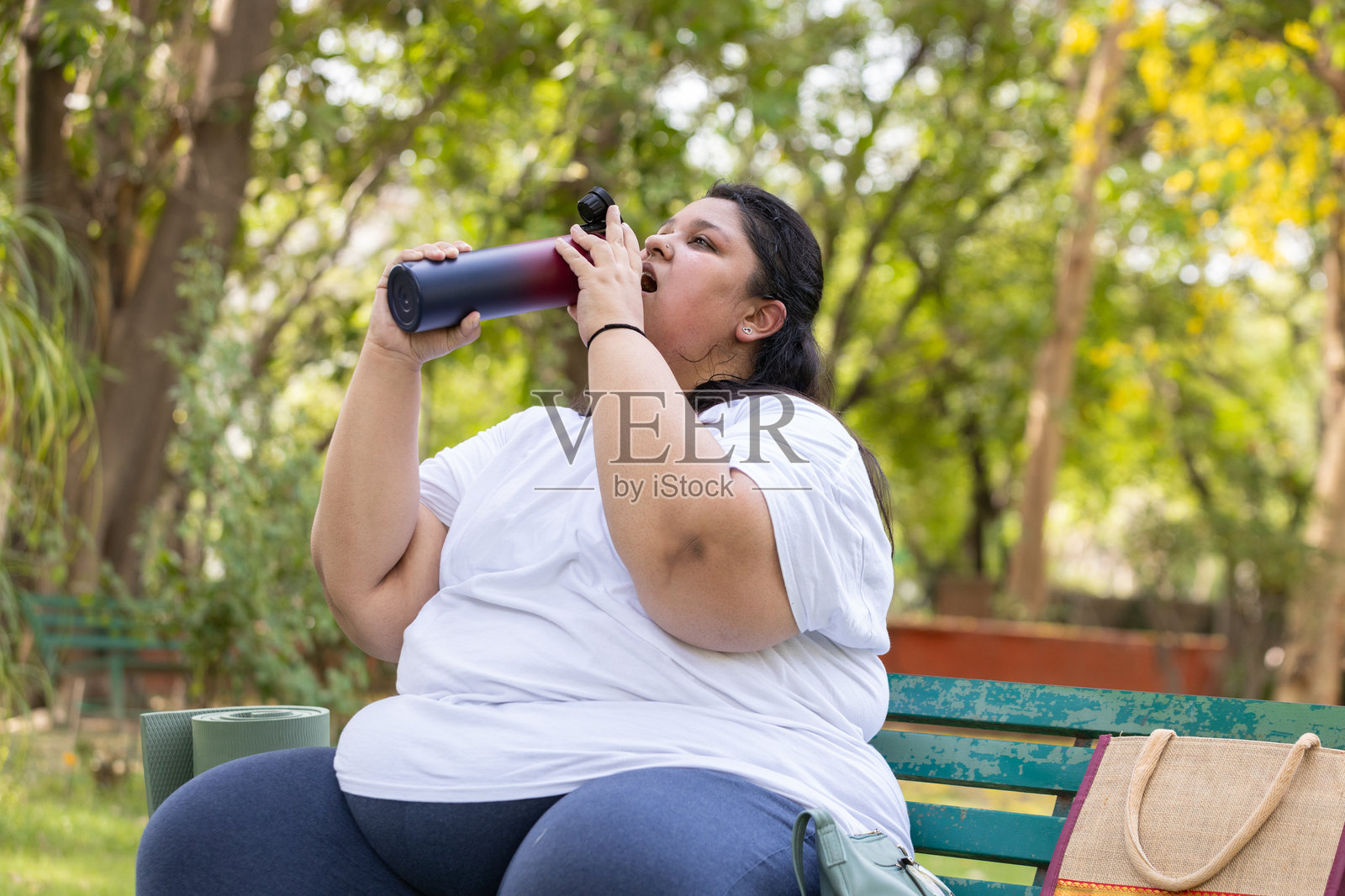 疲惫的印度丰满女性在炎热的夏天喝水。疲惫的超重女士感到口渴。丰满女性在公园户外休息和放松。照片摄影图片