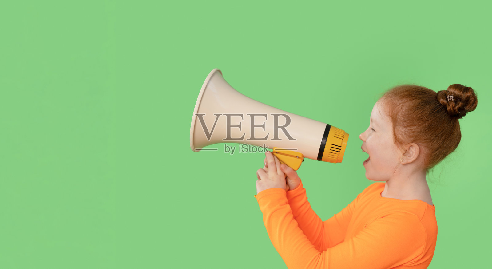 穿着橙色衬衫的孩子在明亮的绿色背景前兴奋地使用扩音器，表达着兴奋和热情。照片摄影图片