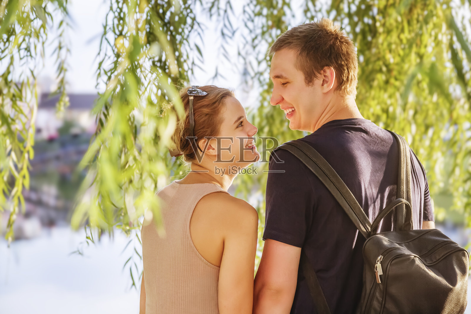 一对年轻情侣站在水边的柳树枝下，相互凝视。照片摄影图片