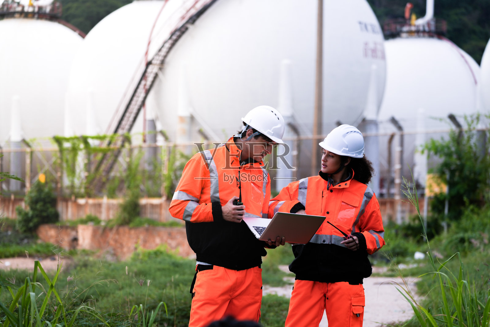 两名穿着高可视安全装备和安全帽的工程师在大型球形气体储存罐前讨论项目计划，手中拿着一台笔记本电脑。照片摄影图片