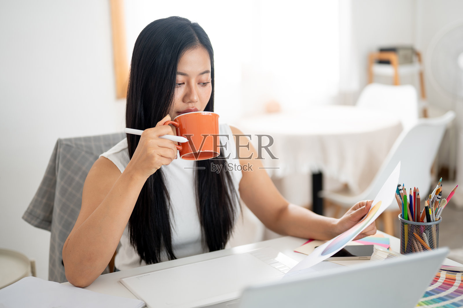 一位亚洲女性正用橙色陶瓷杯喝饮料，同时查看手中的文件。照片摄影图片