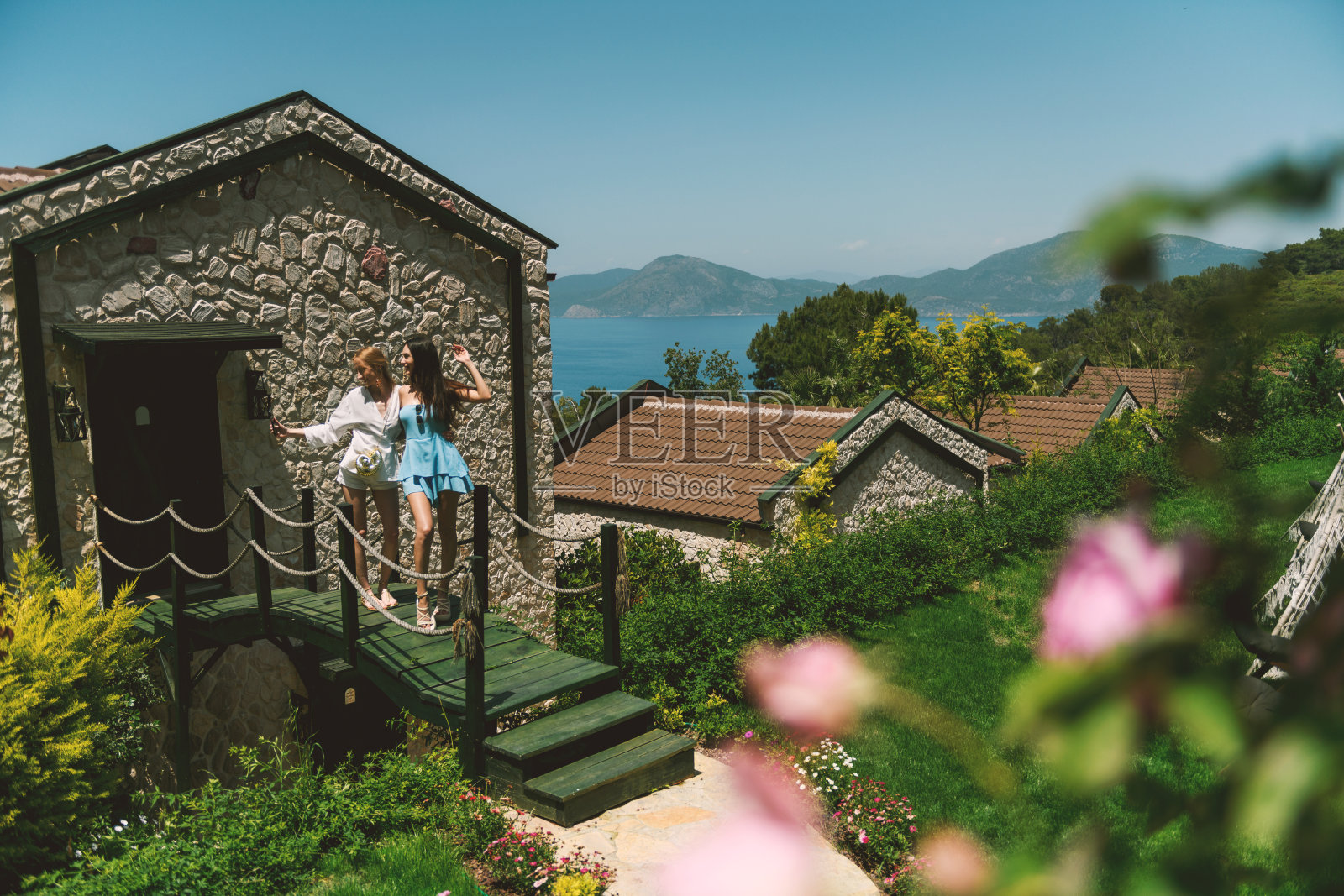 一位穿白色裙子的金发女郎和一位穿蓝色裙子的棕发女郎在酒店花园的美丽桥上自拍。照片摄影图片