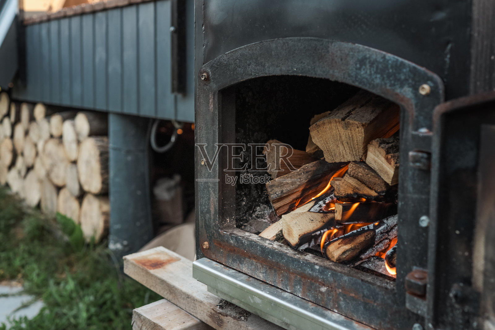 户外金属炉内燃烧着火焰，放置在一座木屋旁边，周围堆放着木柴，现代深色墙板，沐浴在温暖的晚霞中。照片摄影图片