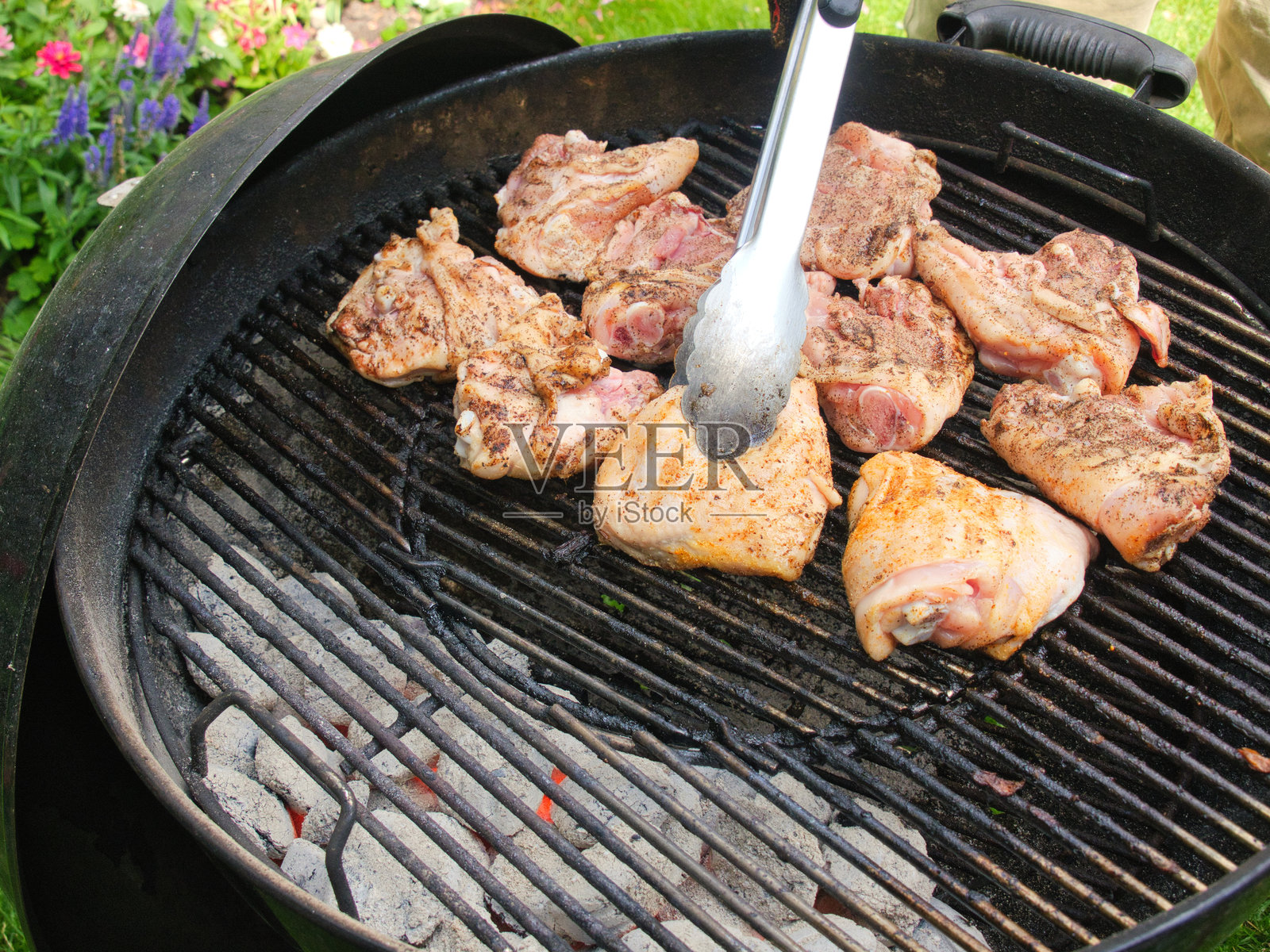 在夏日的户外，使用夹子翻转炭火烧烤锅中正在烹饪的鸡腿。照片摄影图片