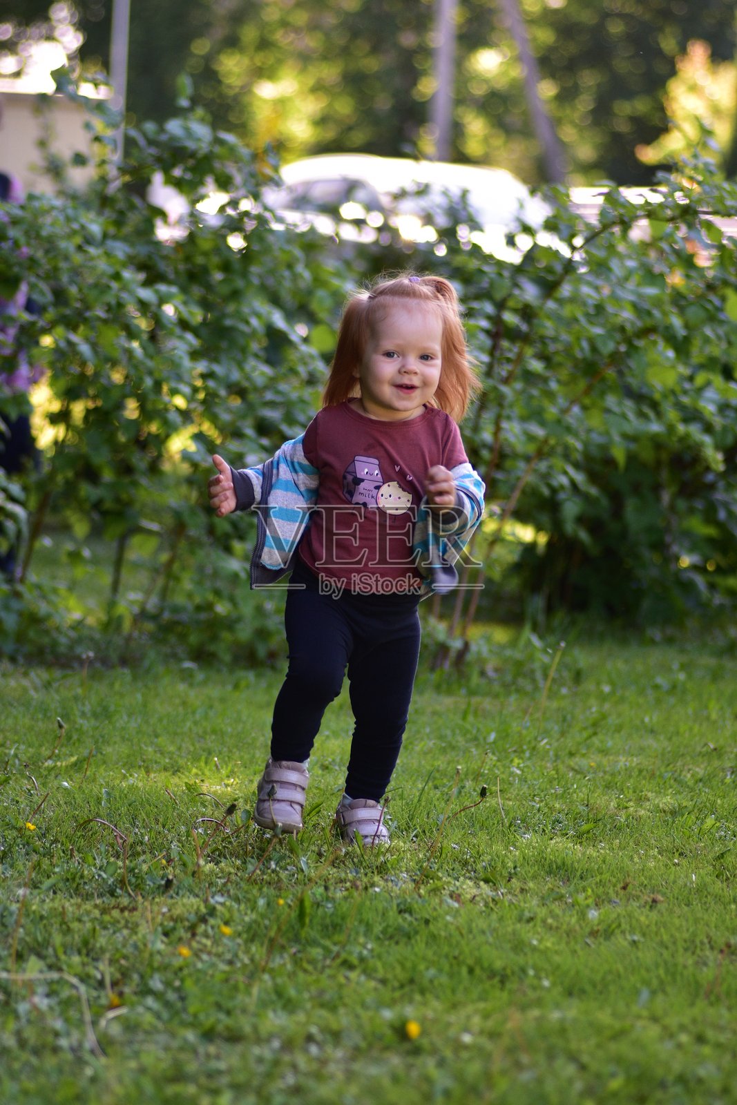 快乐的红发幼女在阳光明媚的日子里户外奔跑，手里拿着一件条纹夹克。在温暖的自然光照射下，生动的绿色花园中捕捉到无忧无虑的童年时刻。照片摄影图片