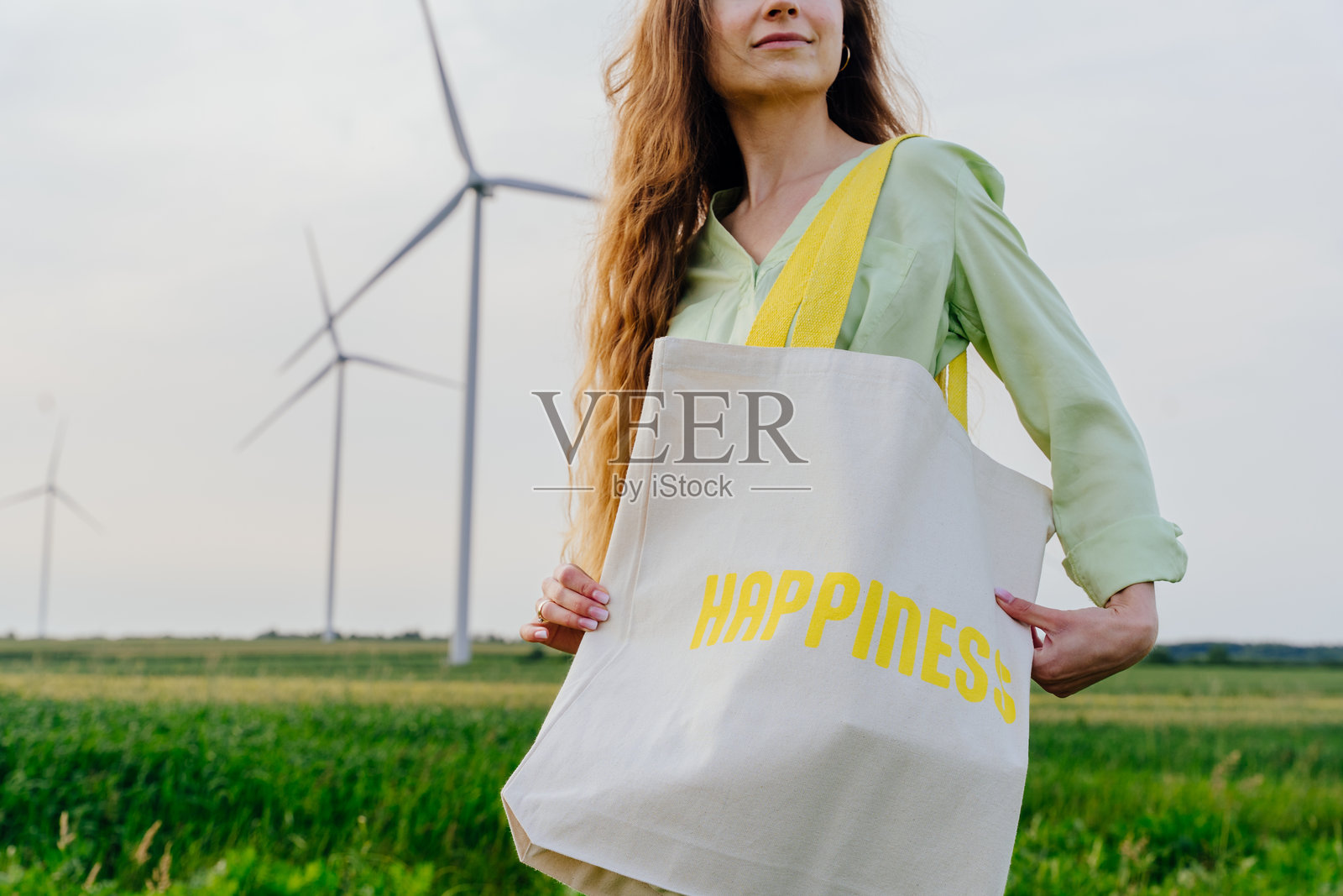 田野中的女性，手提可重复使用的棉布袋，身后是风力发电机照片摄影图片