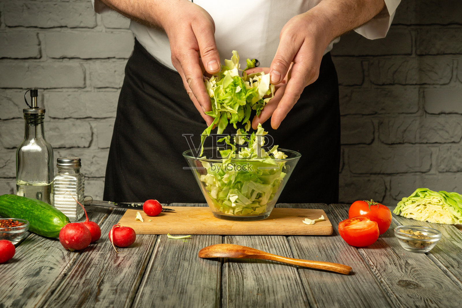 一位厨师在木质台面上用玻璃碗混合新鲜的绿叶蔬菜和其他蔬菜。鲜艳的番茄和黄瓜环绕在他周围，展示了一顿健康的餐食准备。照片摄影图片