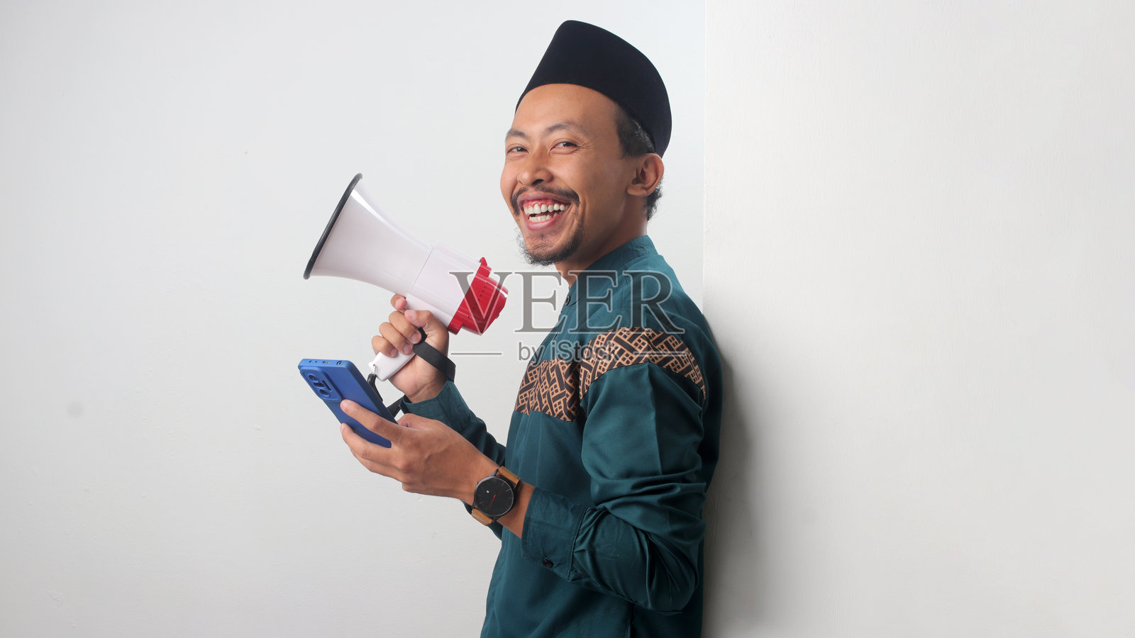 兴奋的亚洲穆斯林男子靠在白墙上，手持扩音器和智能手机照片摄影图片