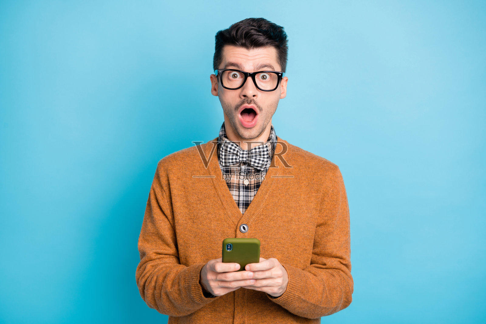 惊讶的男人张嘴，因意外消息而震惊，手持手机，佩戴格子蝴蝶结，背景为蓝色。照片摄影图片