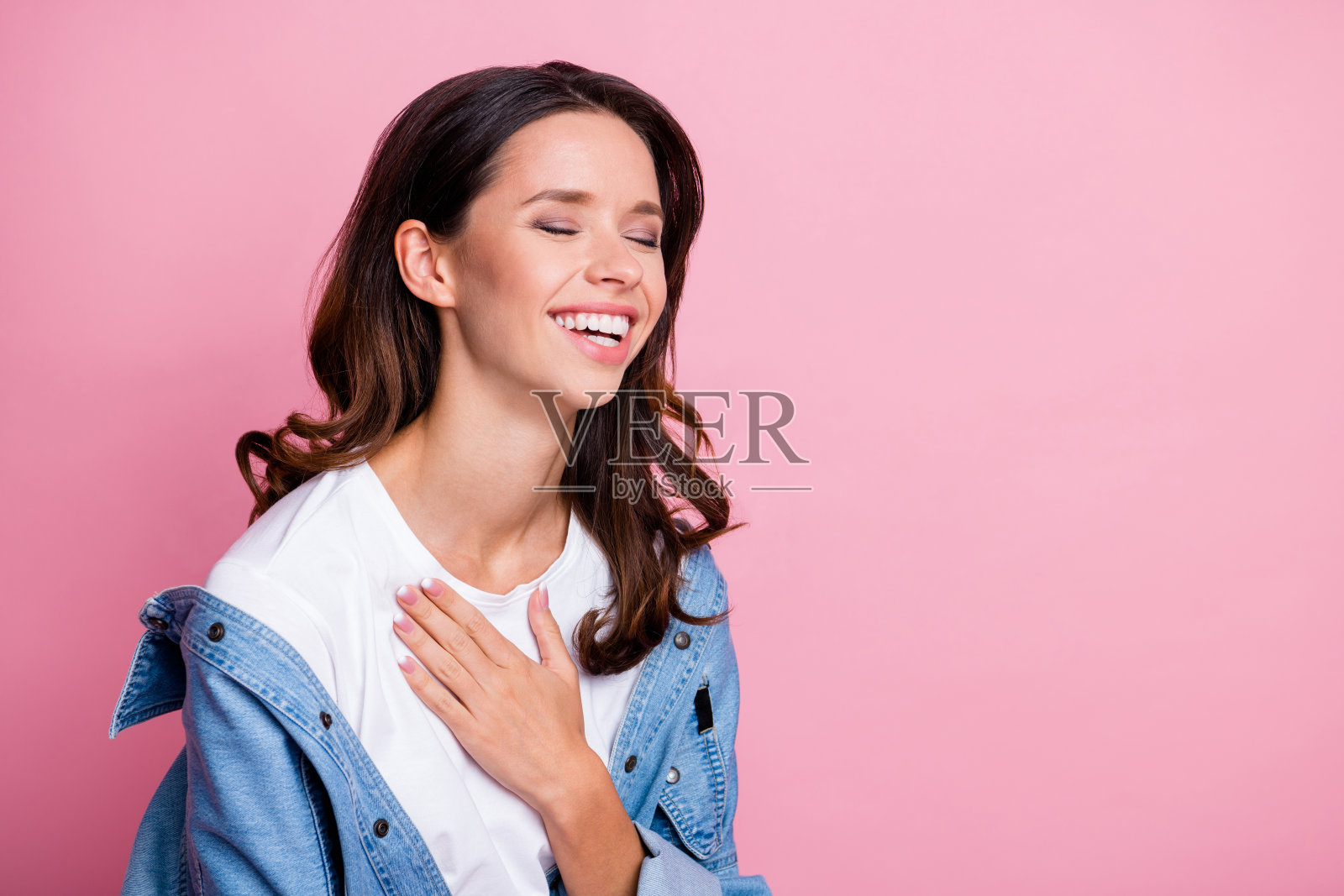 时尚快乐的年轻棕发女性侧面肖像，站在空旷的空间中，笑着讲笑话，背景为柔和的粉色。照片摄影图片