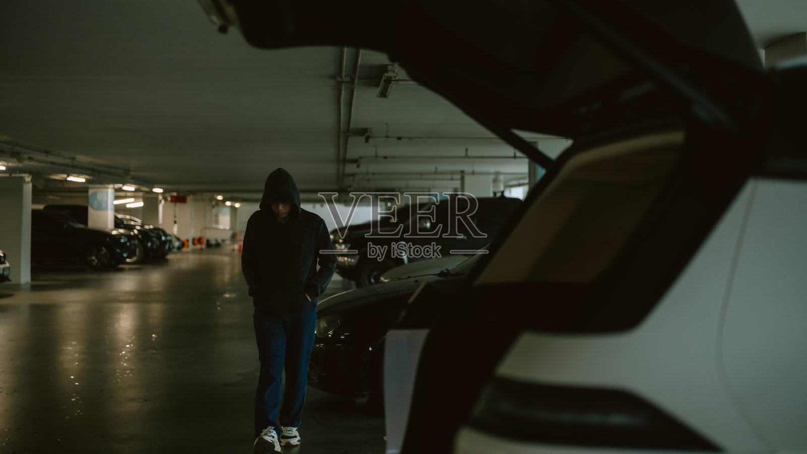 戴着兜帽的人物在昏暗的停车场中走动，周围停满了紧密排列的汽车。照片摄影图片