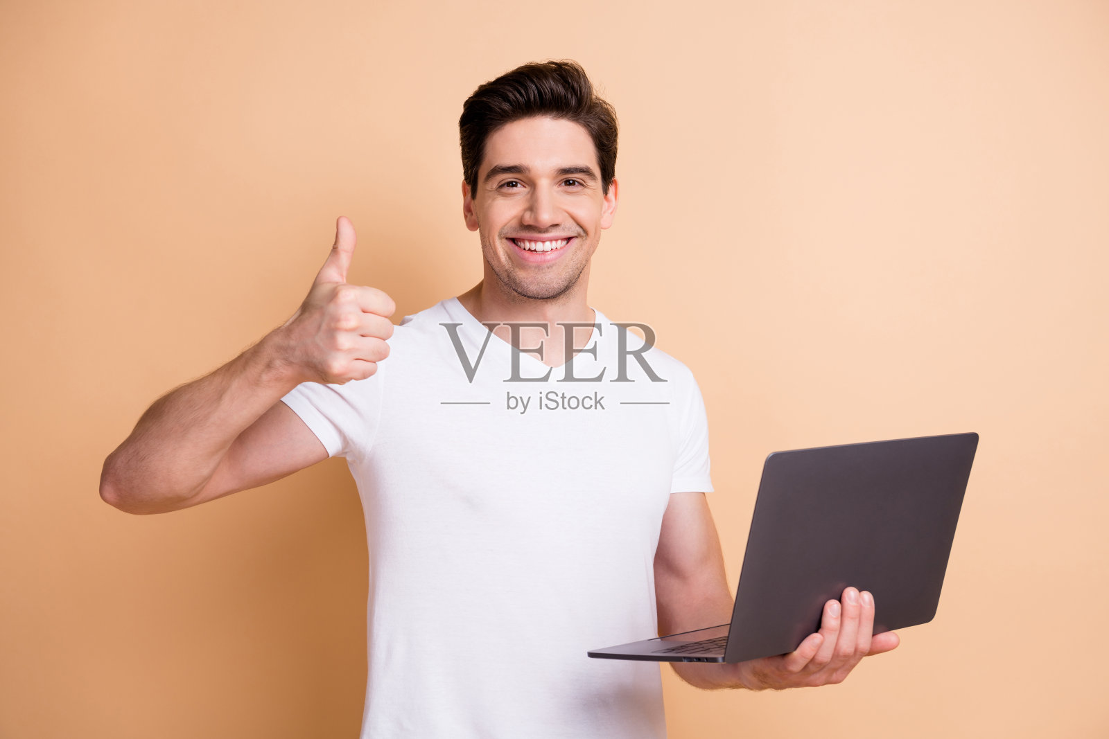 乐观的棕发男士肖像，穿着白色T恤，手持笔记本电脑竖起大拇指，背景为米色。照片摄影图片