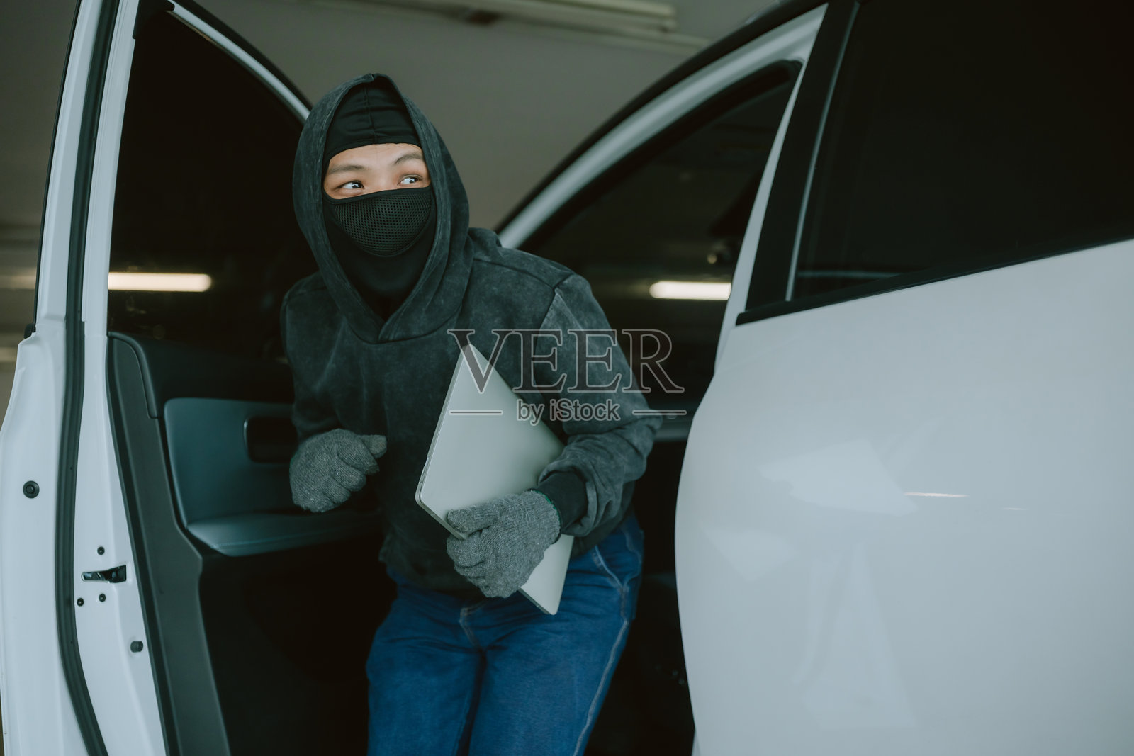 身穿深色衣物的人在昏暗的停车场中悄悄走出汽车，手中拿着笔记本电脑，正值傍晚时分。照片摄影图片