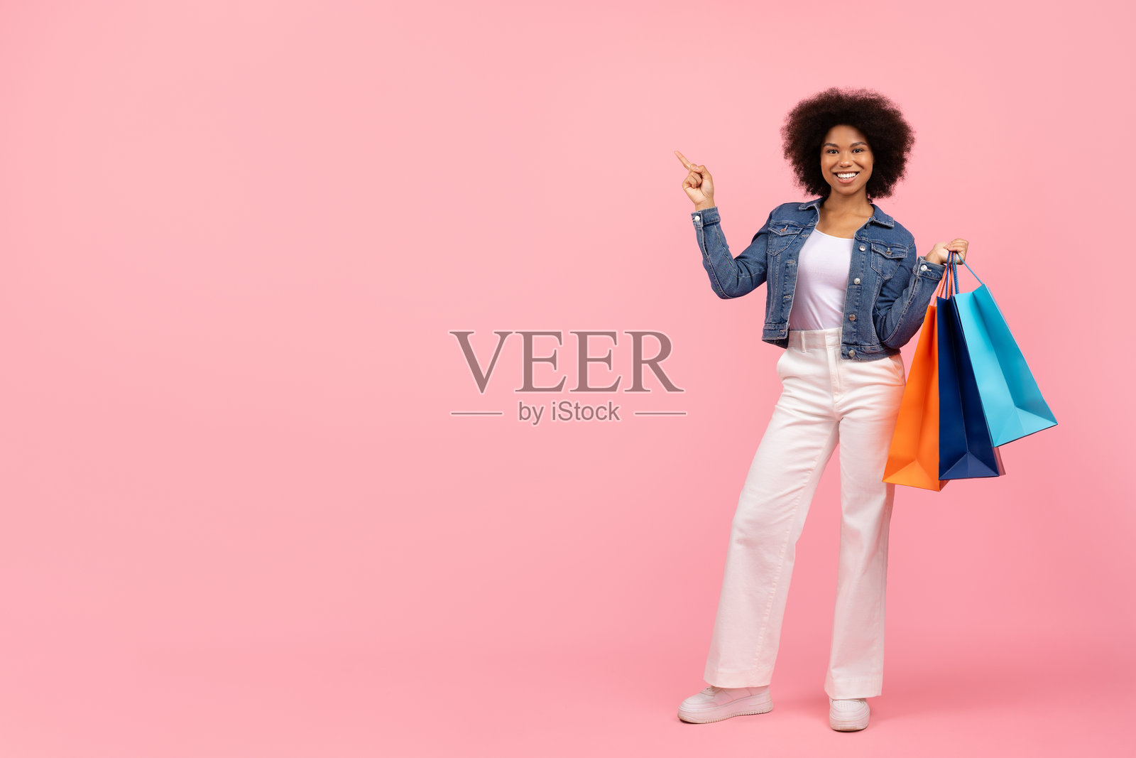 时尚的年轻黑人女性微笑着指向一旁，手里提着购物袋。照片摄影图片