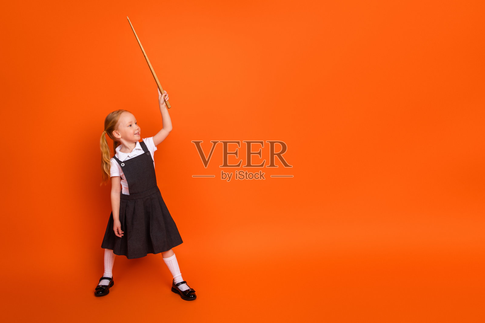 可爱的年轻女学生在橙色背景下手持指示棒，面露喜悦，身穿校服，扎着马尾辫。照片摄影图片