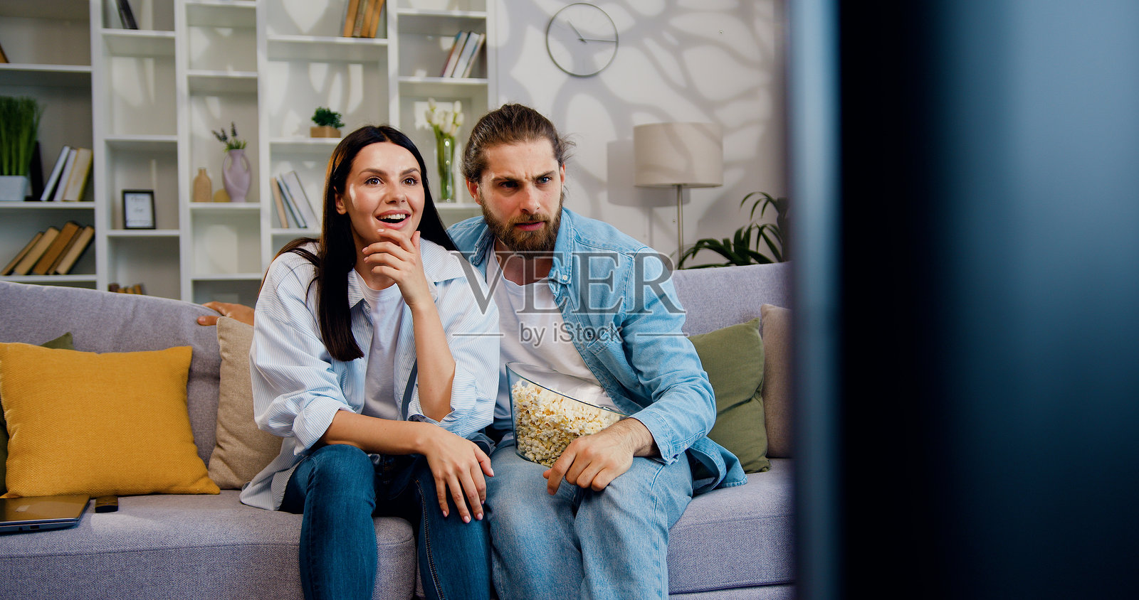 家庭氛围的概念，吸引人的无忧无虑的情侣男士和女士坐在沙发上，面前是电视，吃着爆米花，交谈并在沙发上跳跃。照片摄影图片