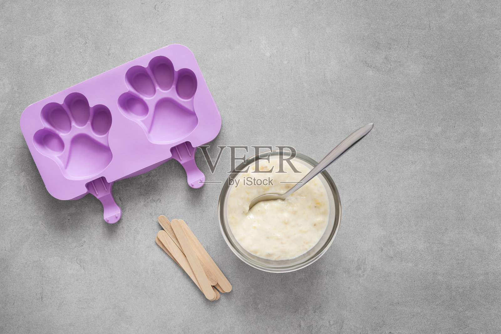 紫色硅胶爪形模具，碗中盛着奶油混合物，灰色混凝土背景上的木棒照片摄影图片