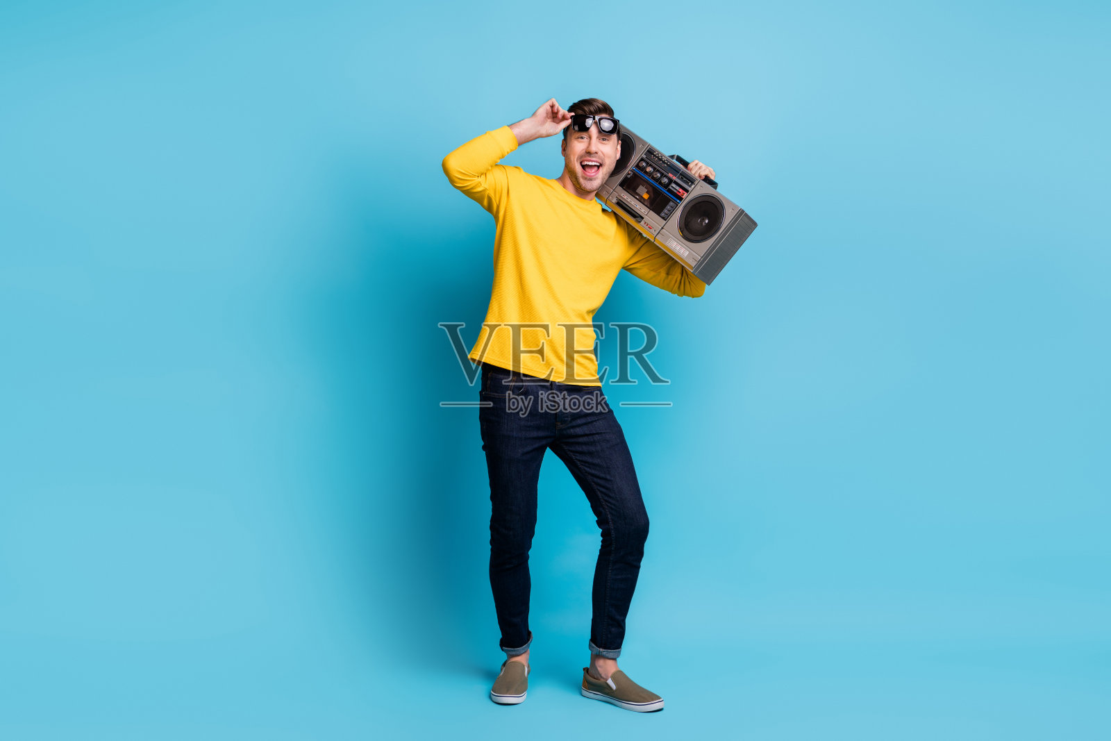 全身照，快乐的年轻人手持太阳镜和音响，背景为蓝色。照片摄影图片
