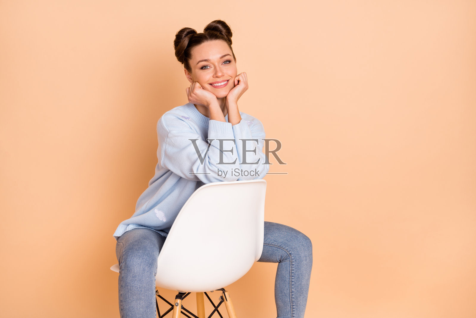 乐观的棕发女士坐着，双手托着脸，穿着毛衣和牛仔裤，背景为柔和的米色。照片摄影图片