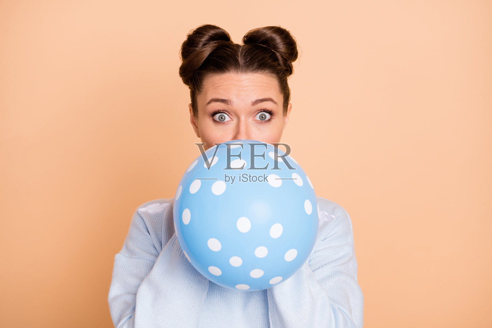 乐观的棕发女士穿着蓝色毛衣，吹气球，背景为柔和的米色。照片摄影图片