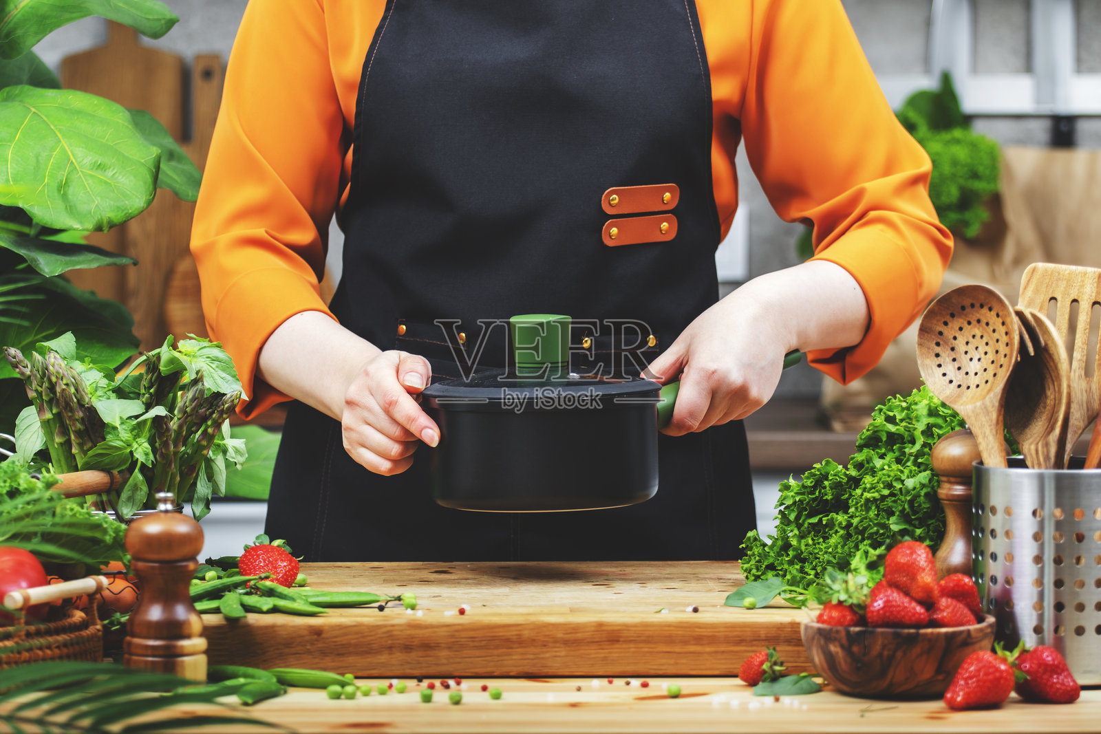 穿围裙的女厨师手持平底锅，准备在一个装满健康食材的整洁厨房中烹饪新鲜蔬菜。照片摄影图片
