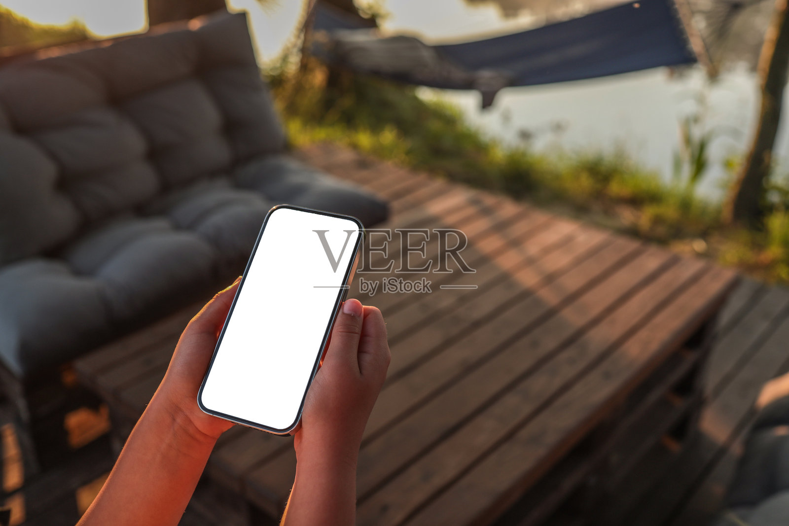 手握空白屏幕智能手机，坐在湖边的木质露台上，日出时分。背景中的吊床和舒适的户外家具营造出轻松的夏日氛围，适合用于样机展示。照片摄影图片