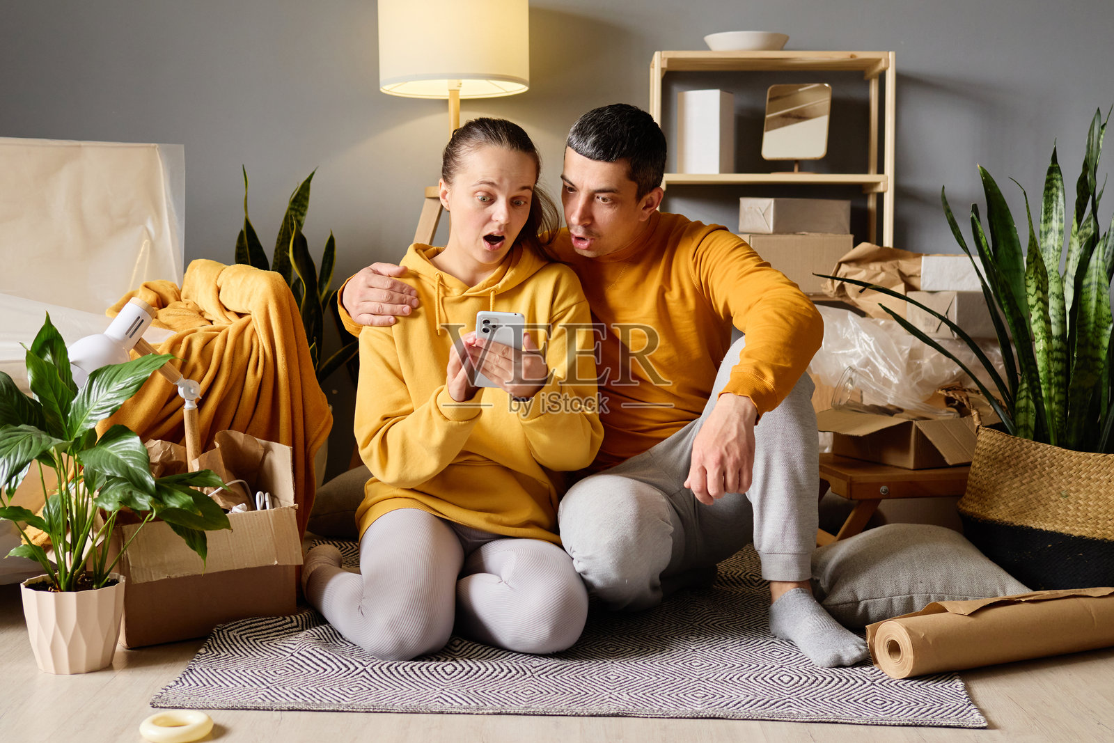 一对惊讶的情侣跪在地板上，周围是移动箱子，女人手中拿着一部智能手机。照片摄影图片