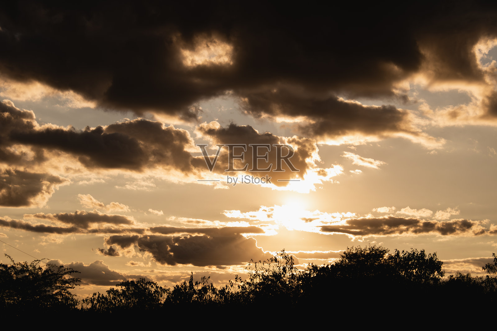 阳光在金色时刻透过多层云彩照耀，树木的轮廓在其后显现。照片摄影图片