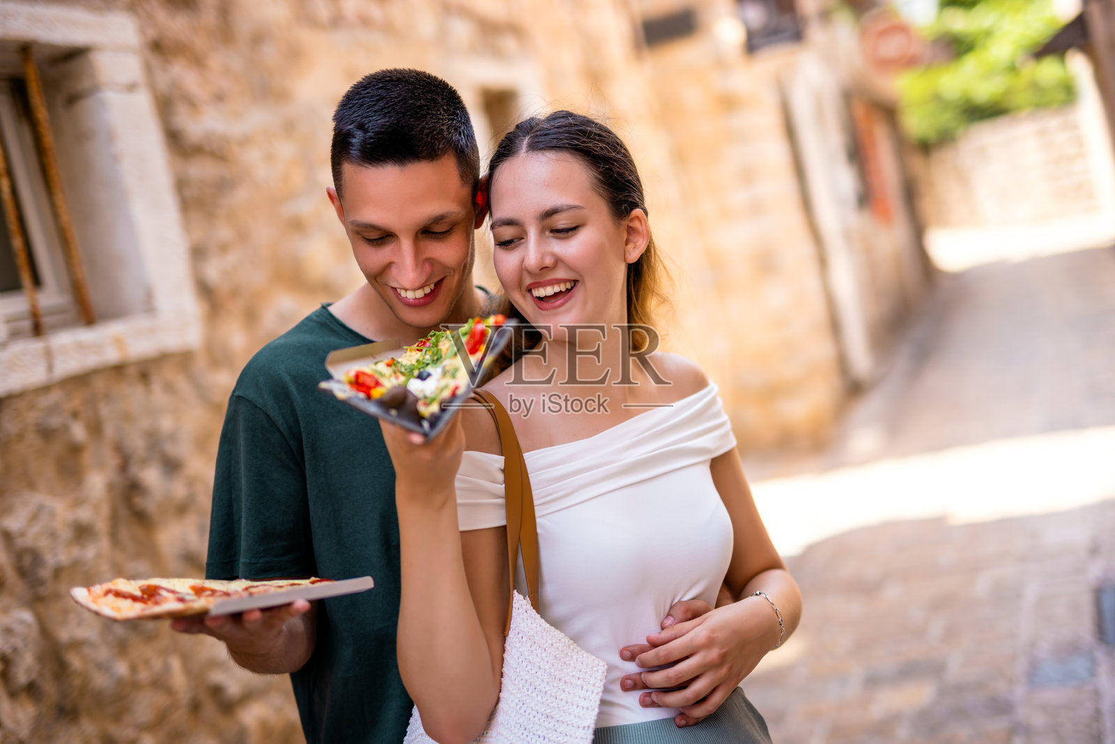 男朋友从后面拥抱女朋友，她在夏季假期中给他喂披萨，地点在老城区。照片摄影图片