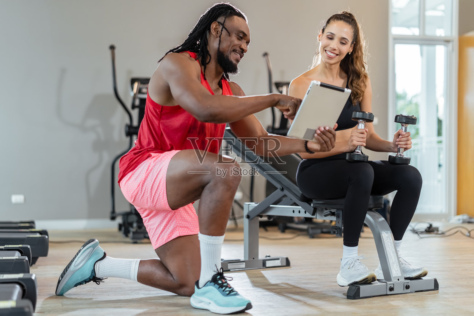 一男一女在健身房。男子指着一台平板电脑，女子手里拿着两个哑铃。照片摄影图片