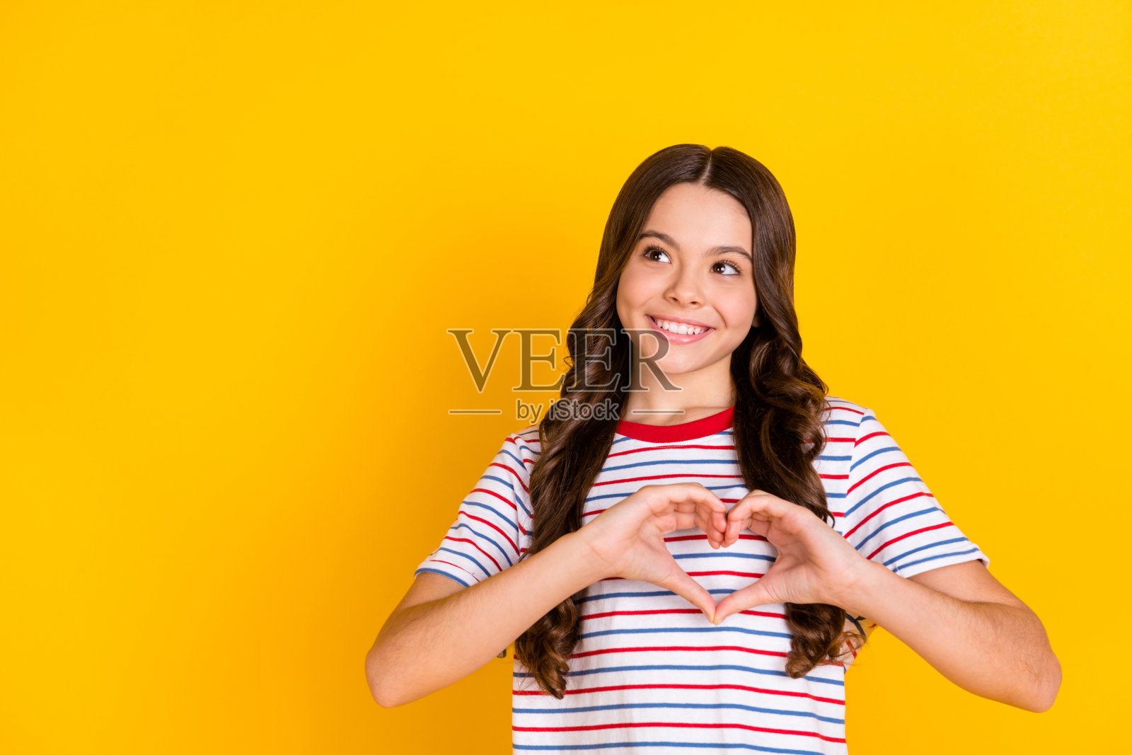 年轻女孩开心地微笑，梦幻般地看着空旷的空间，手指比出心形，表达爱意，背景为黄色。照片摄影图片