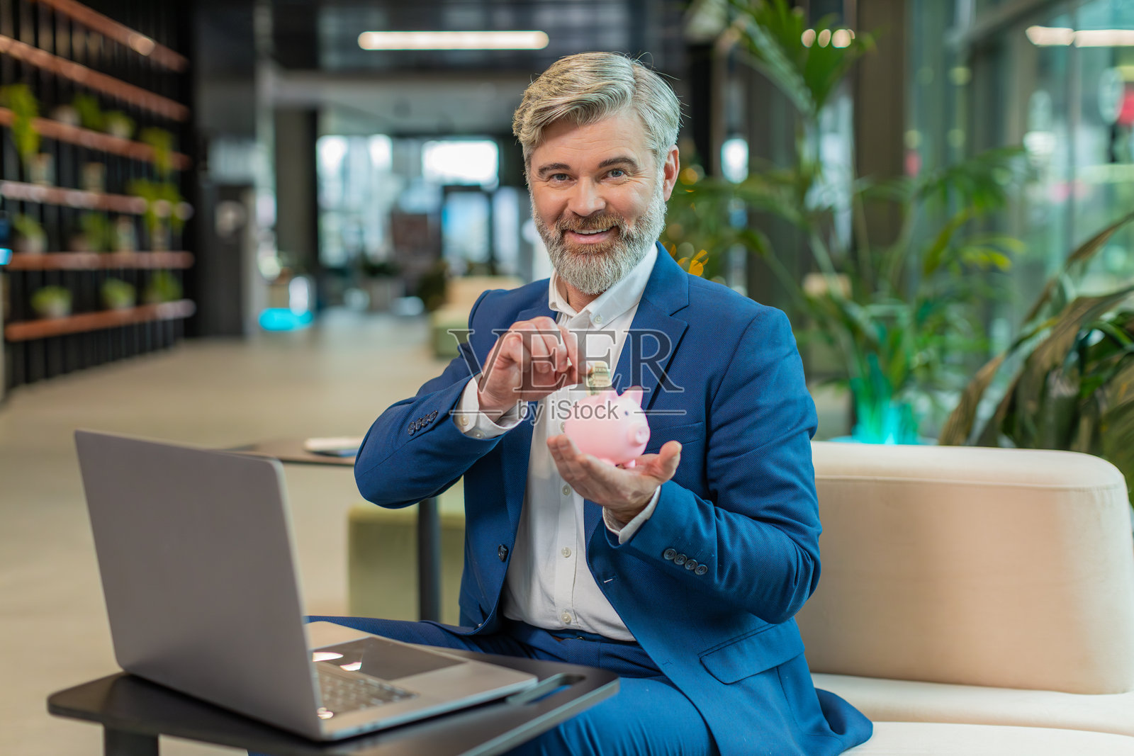 快乐的成熟商人在办公室大堂用粉色储蓄罐存钱，把美元放进去照片摄影图片