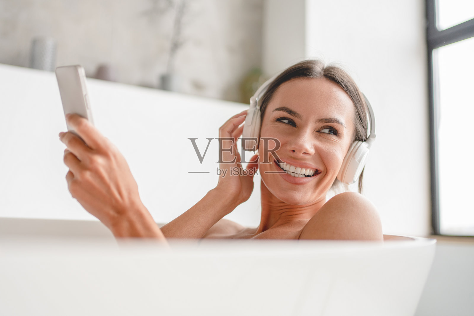 女性在现代室内浴室放松，穿着无线耳机，手持手机，享受洗澡的乐趣。愉快的女性闭着眼睛在家中享受沐浴时光。照片摄影图片