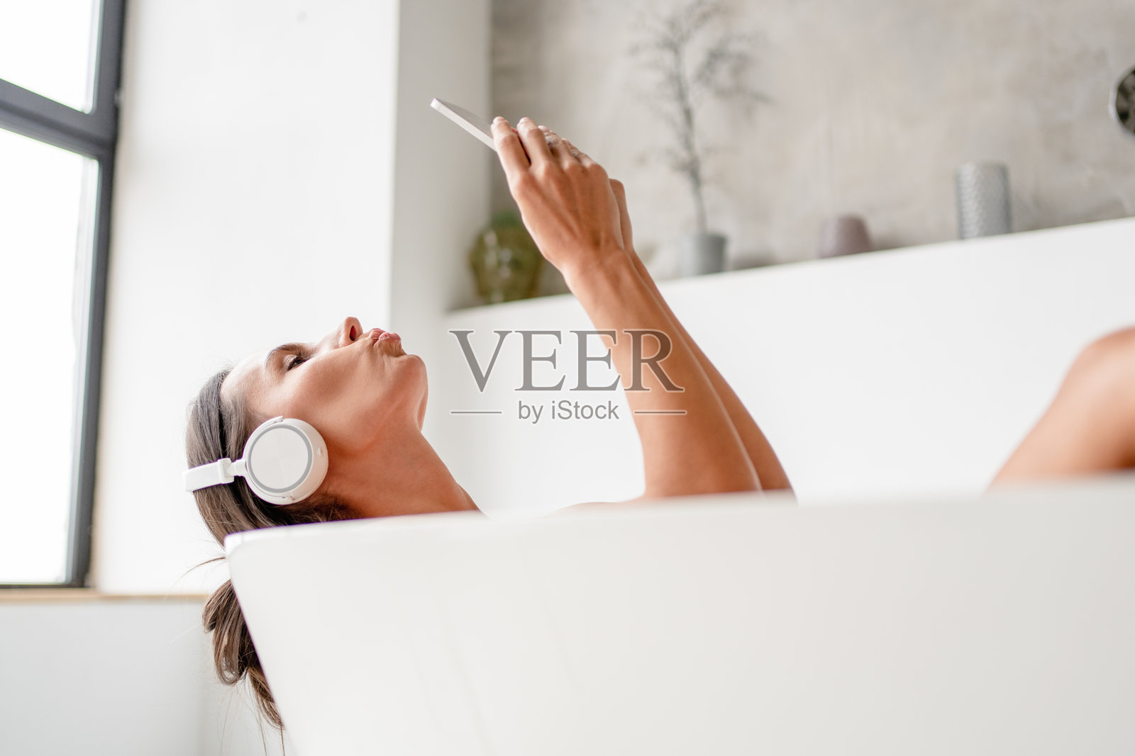 年轻女士在泡沫浴缸中放松，戴着无线耳机听音乐，手中拿着智能手机自拍，悠闲地在家浴室中休息。放松的女性闭着眼睛享受沐浴时光。照片摄影图片