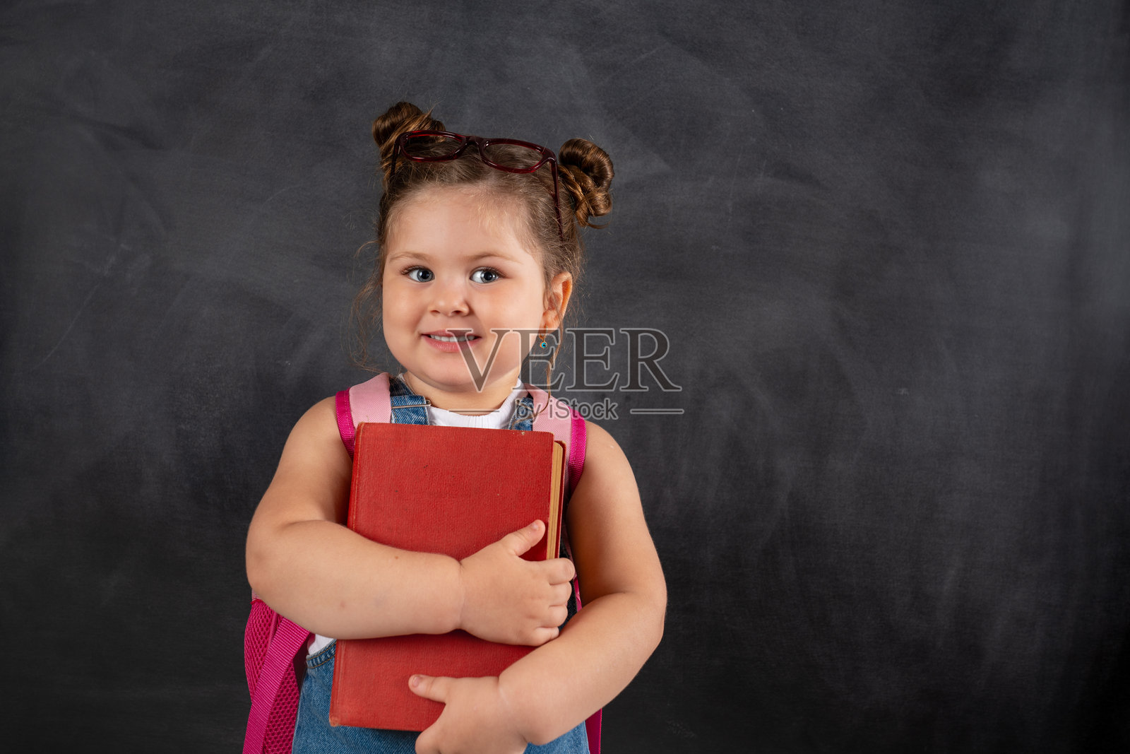 可爱的小女孩在黑色黑板前微笑着，手里拿着一本书。照片摄影图片