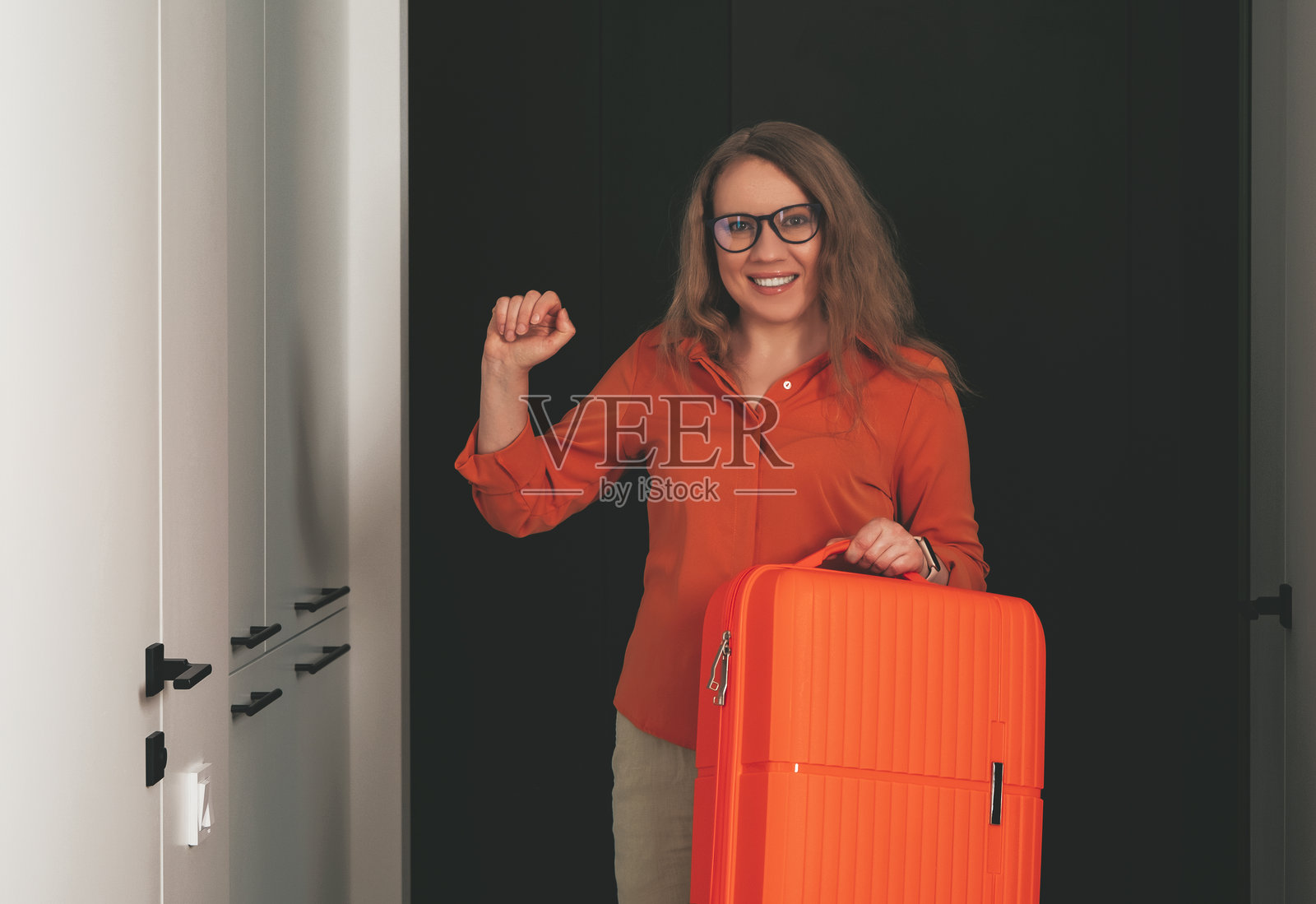 快乐的金发女人在时尚公寓中准备夏季假期，旁边有一个鲜艳的橙色行李箱。照片摄影图片