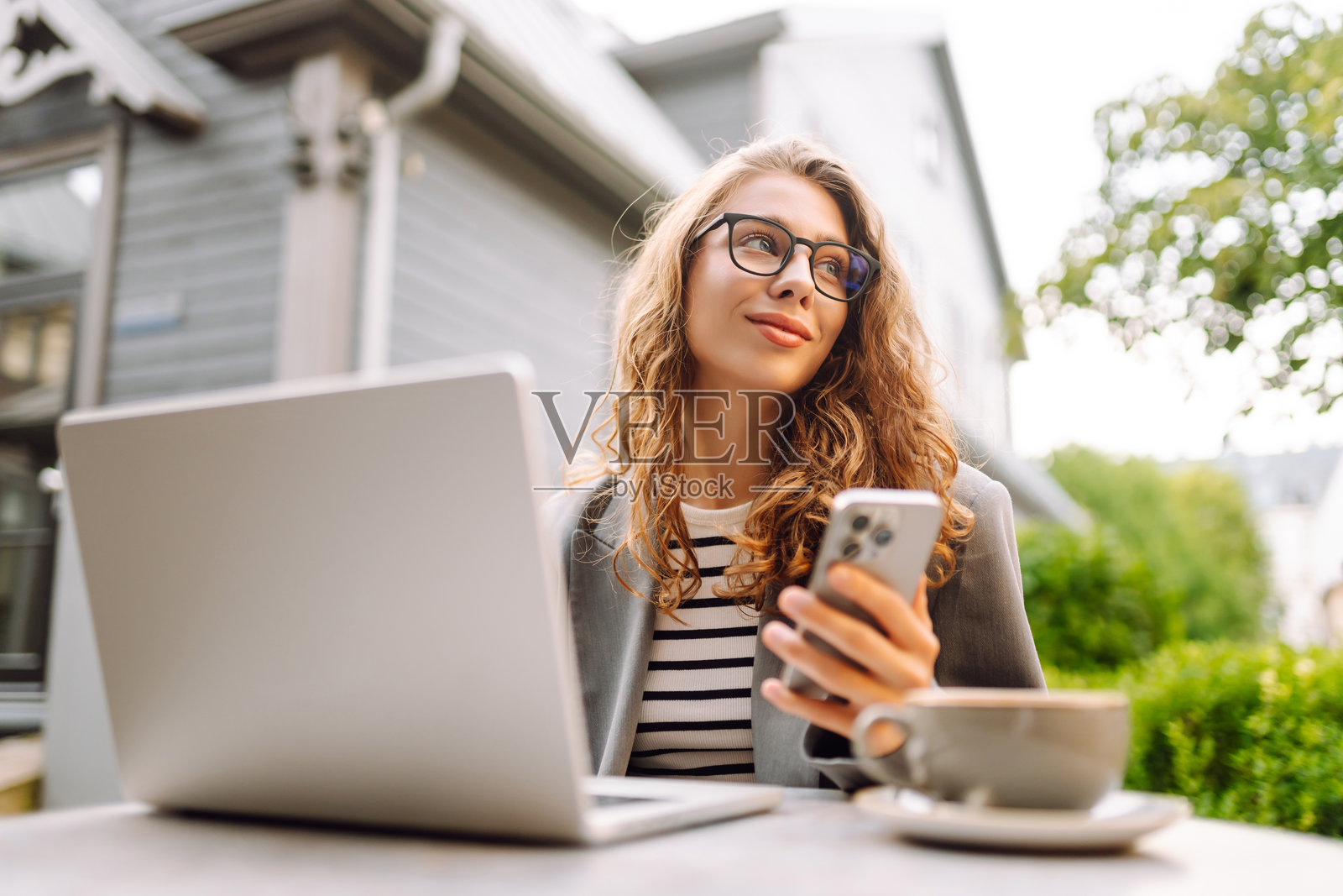 一位年轻女性坐在咖啡馆阳台的桌子旁，手中拿着笔记本电脑和手机。照片摄影图片