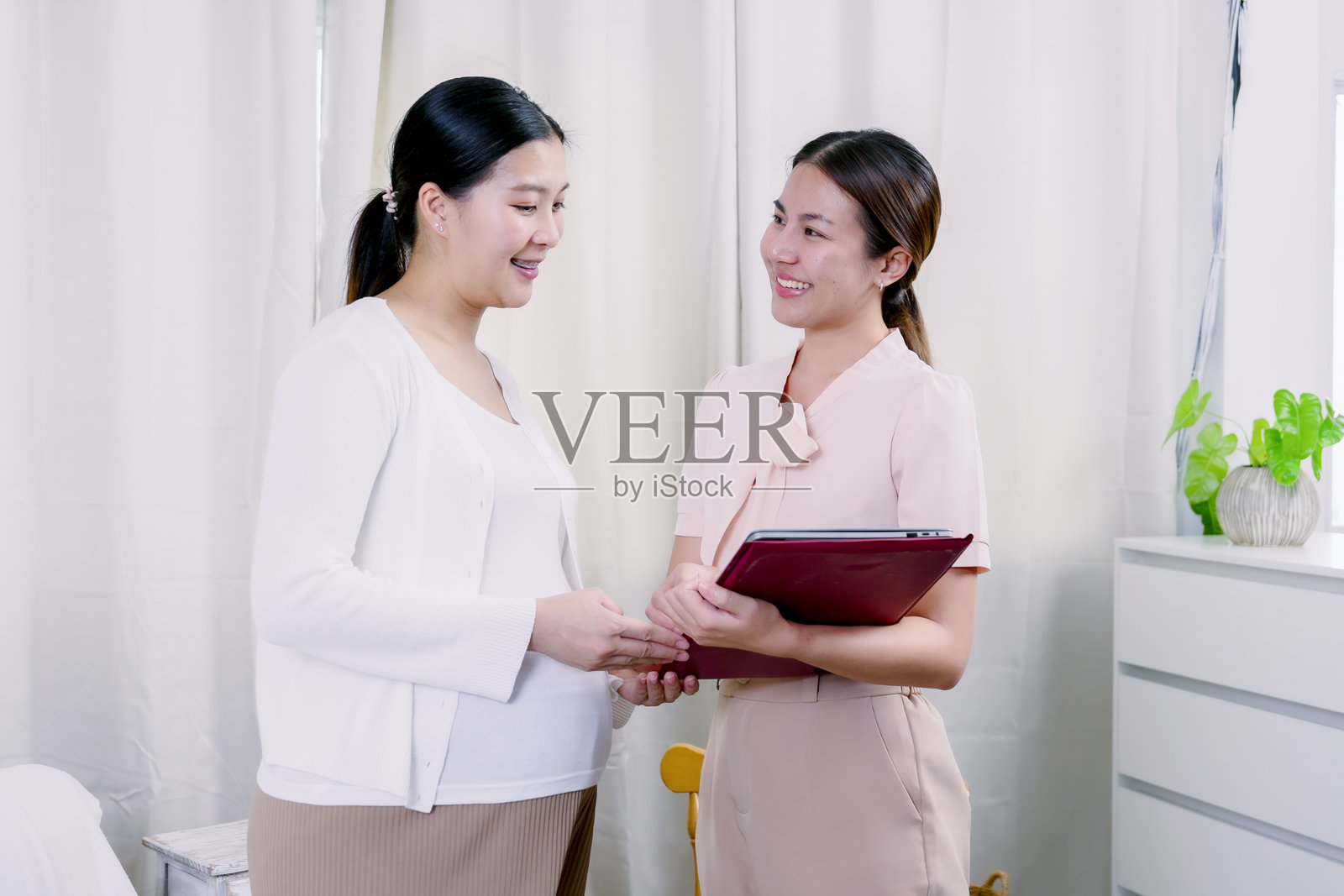 亚洲孕妇和女性代理微笑着站在一起，手持保险文件，展现出在温馨友好的家庭环境中安排新生儿保险计划后的满意和信任。照片摄影图片