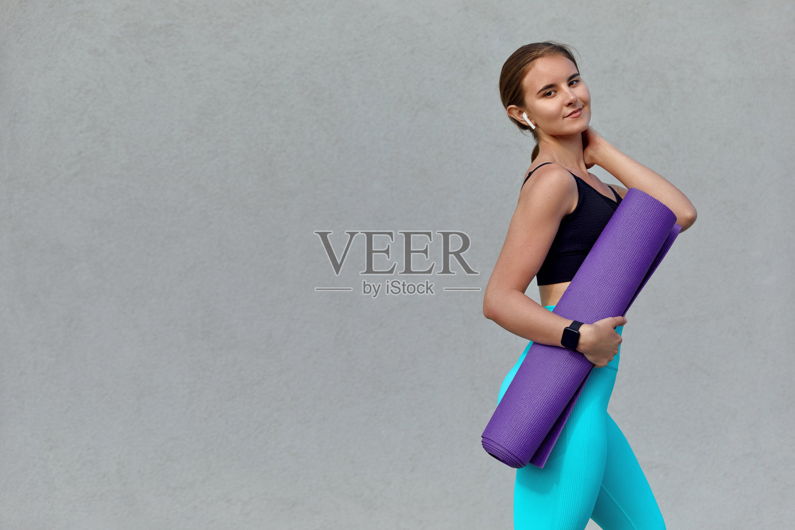 年轻的运动女性自信地走在户外，手里拿着瑜伽垫。她穿着健身服，散发着健康和快乐，体现了对健康和放松的承诺。照片摄影图片