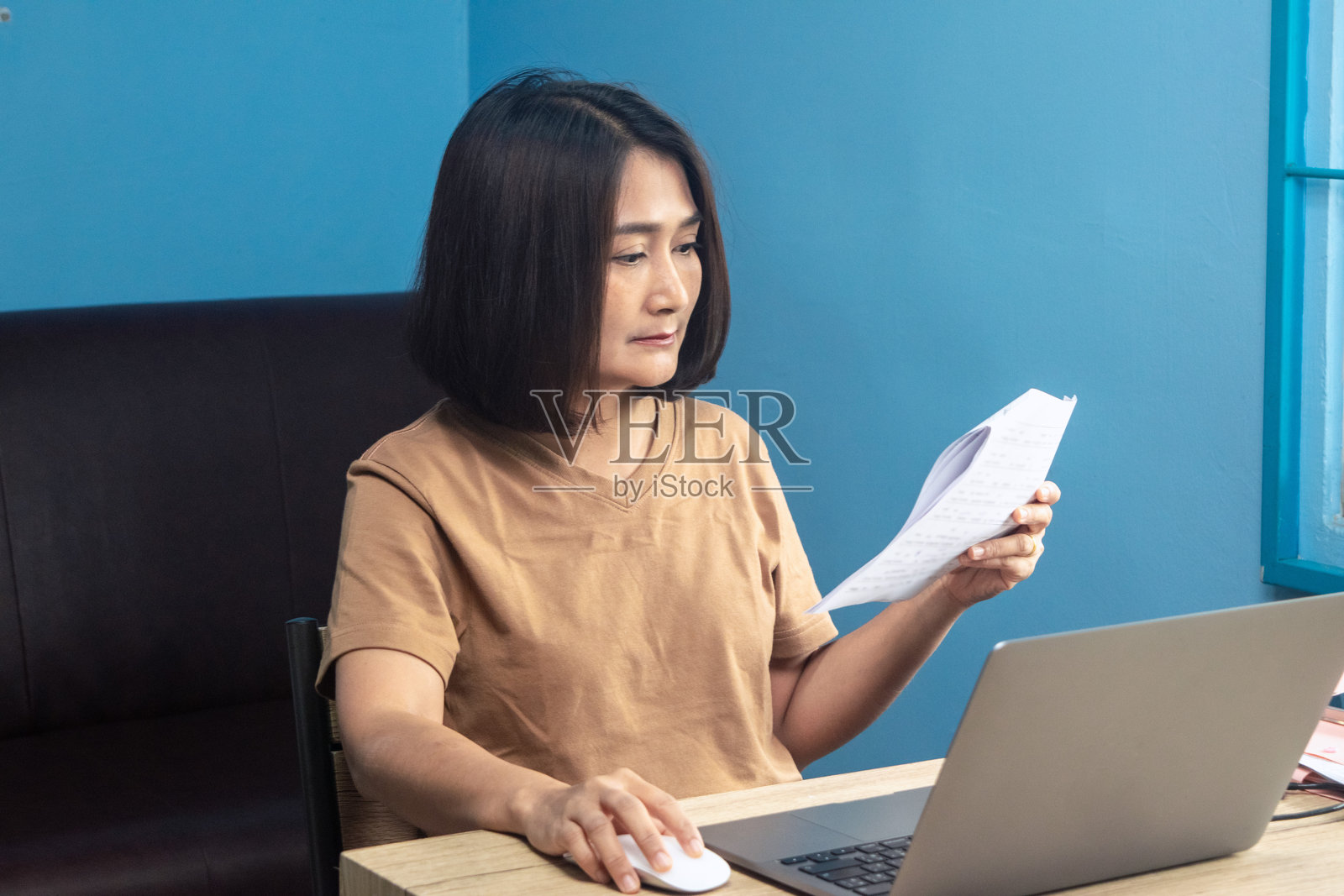 女性坐在椅子上，居家办公，手持纸质文件，使用桌上的笔记本电脑，在线学习提升商业技能，度假生活方式概念照片摄影图片