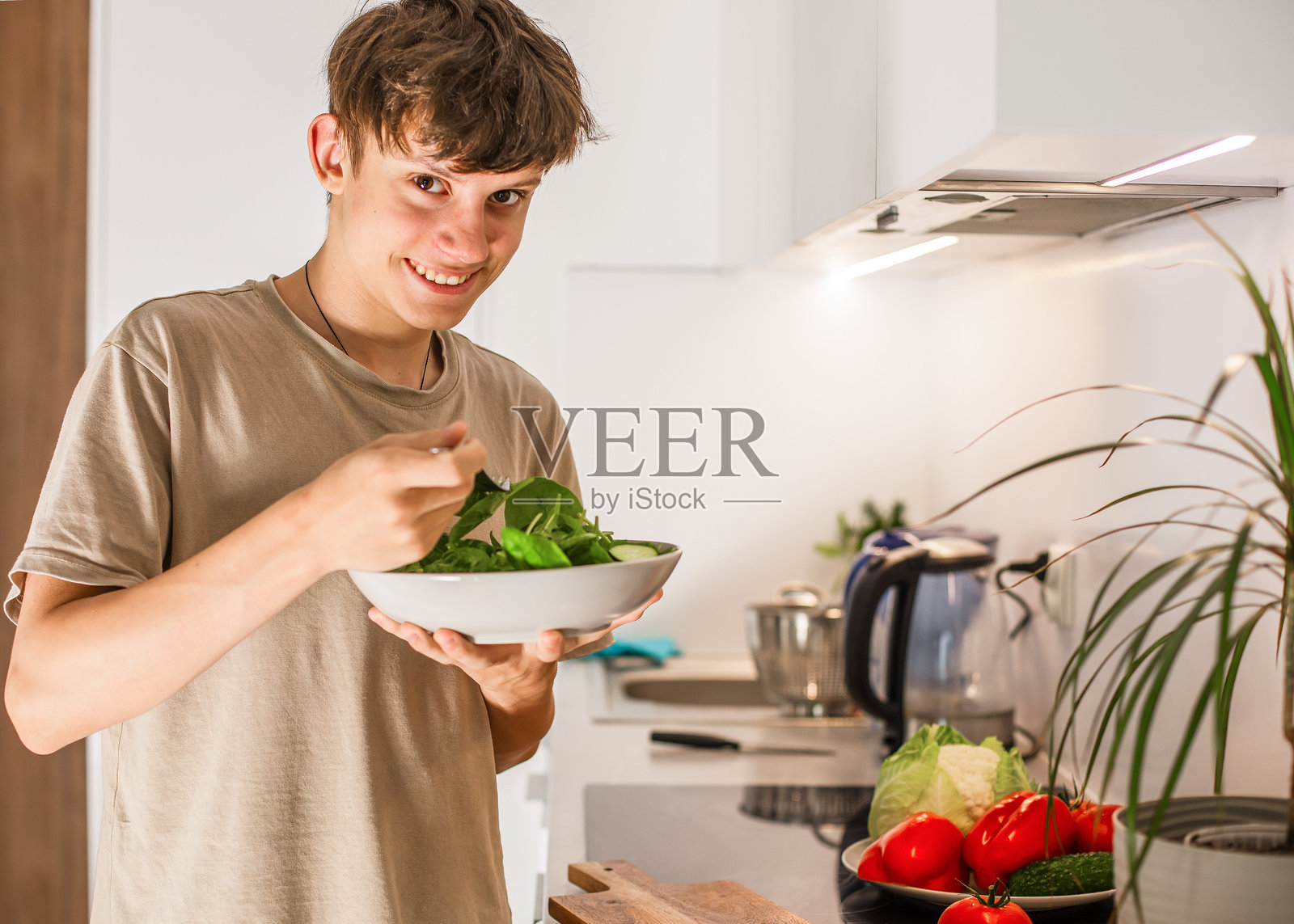 快乐的少年在厨房里准备了一份新鲜蔬菜的素食沙拉，并愉快地享用它。照片摄影图片
