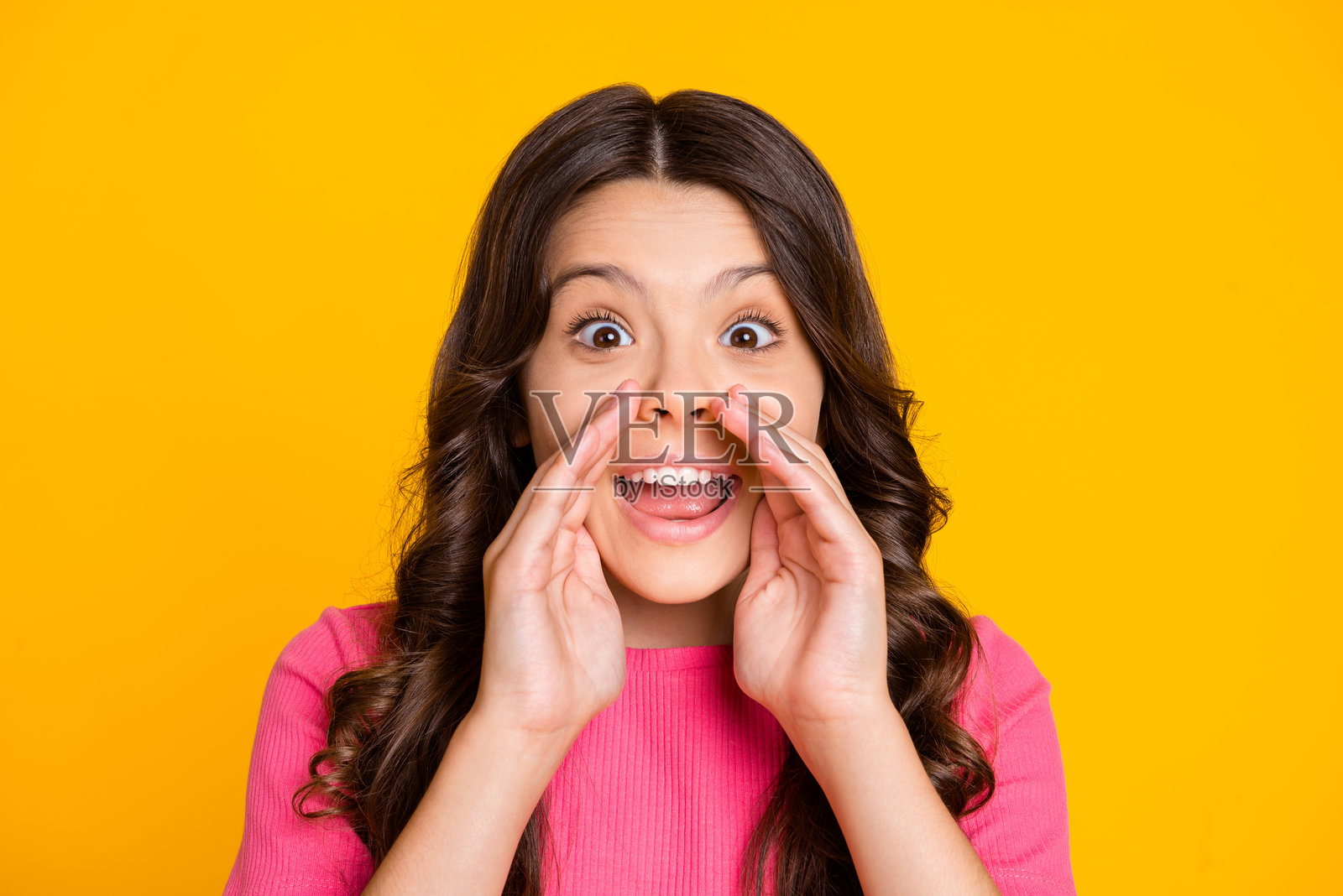 快乐兴奋的女孩双手捂嘴，低声耳语秘密，背景为明亮的黄色。照片摄影图片